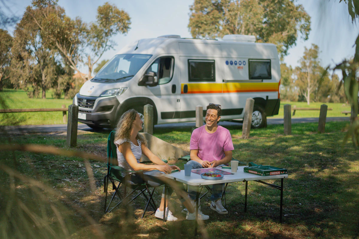 Two people sitting at a picnic table by a campervan, enjoying a sunny day in a green park.