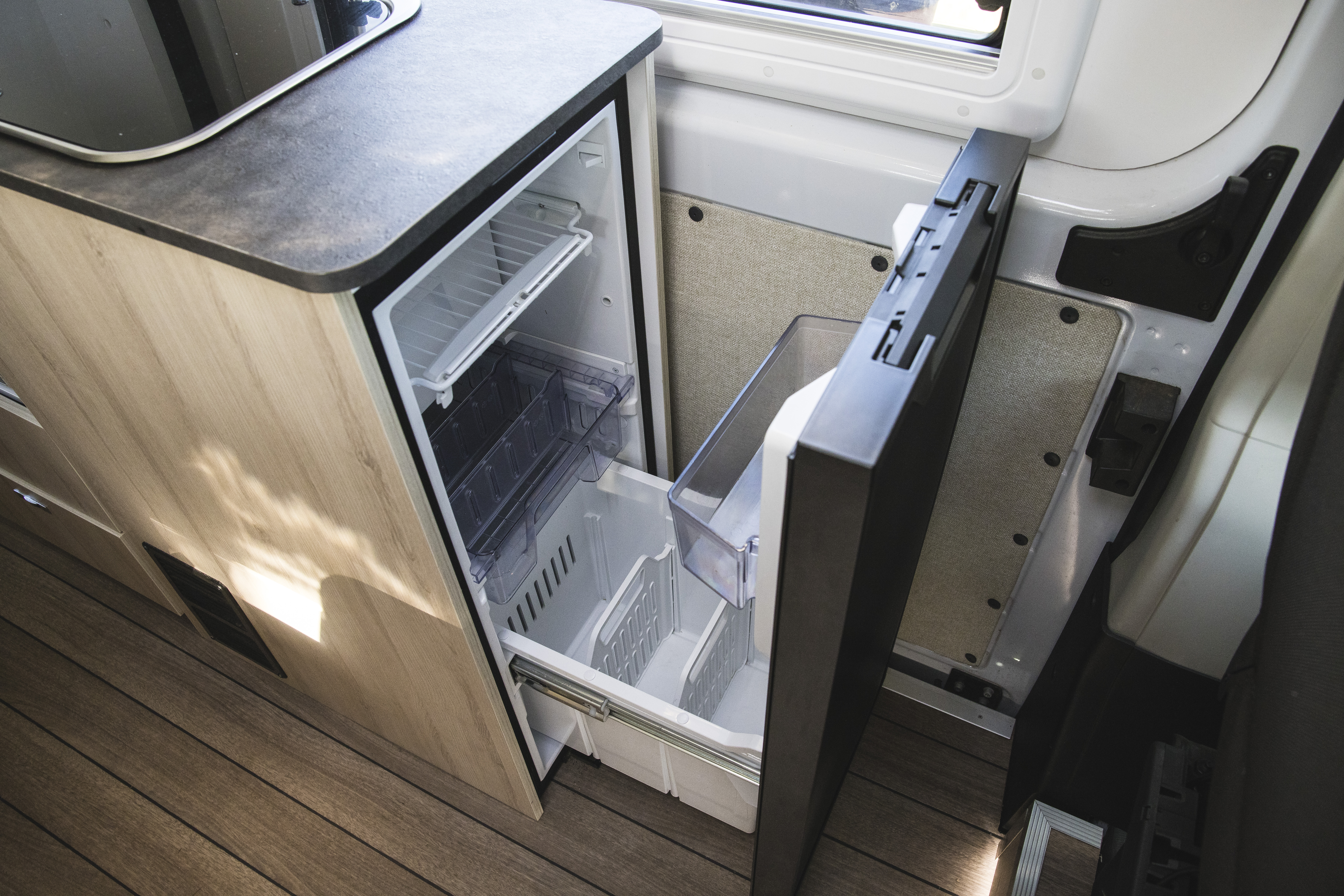 Compact campervan kitchen with an open refrigerator and freezer, wood cabinetry, and natural light from a nearby window.