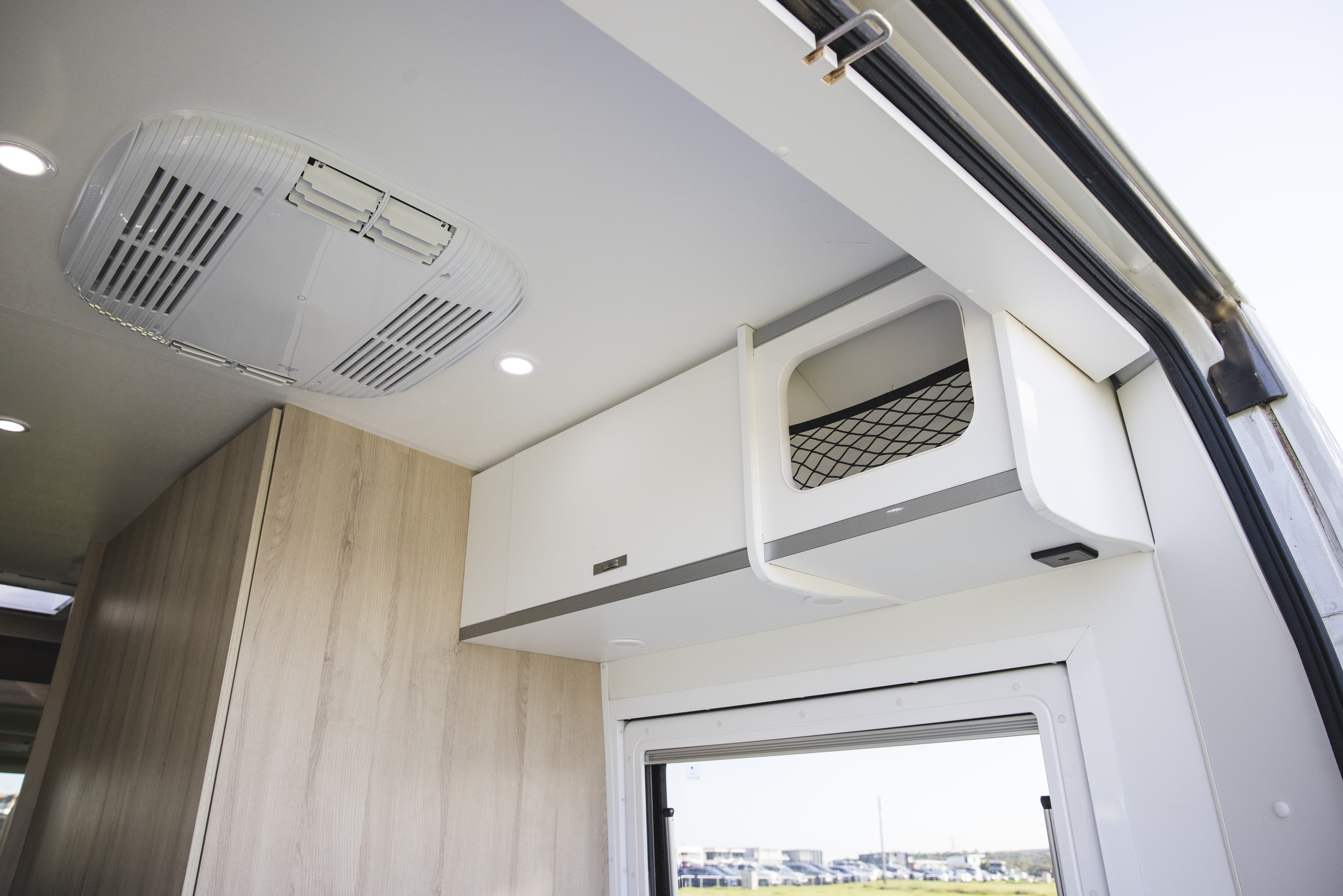 Interior view of a campervan showing built-in overhead storage, wood paneling, air conditioning, and a window.