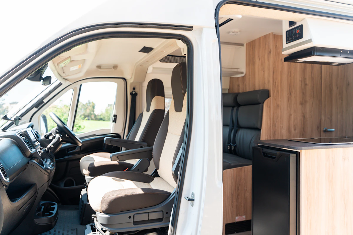 Interior view of a camper van showing driver seats, kitchenette with fridge, and a small seating area.
