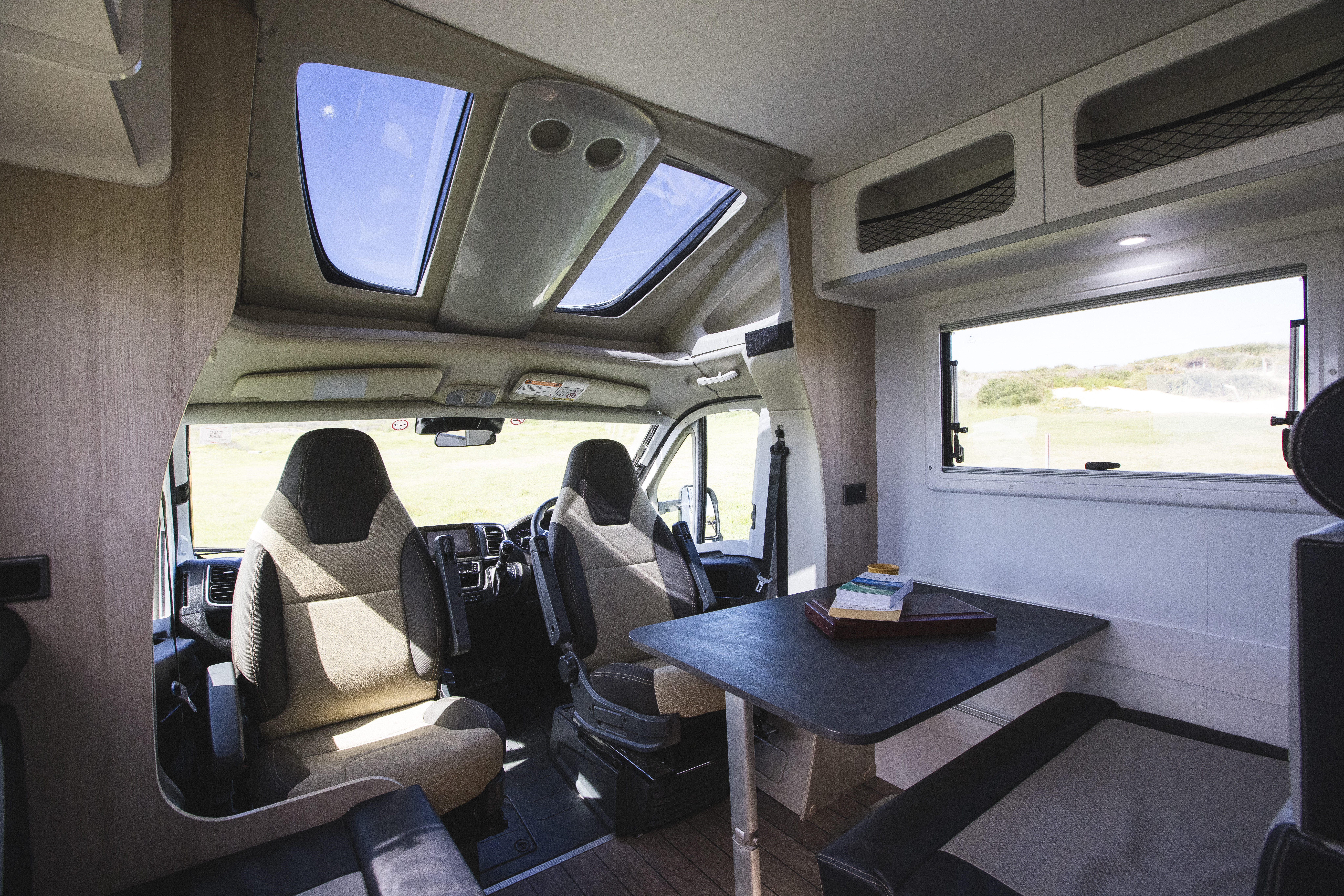 Modern campervan interior with two swivel seats, a dining table, skylights, and a window with scenic outdoor view.