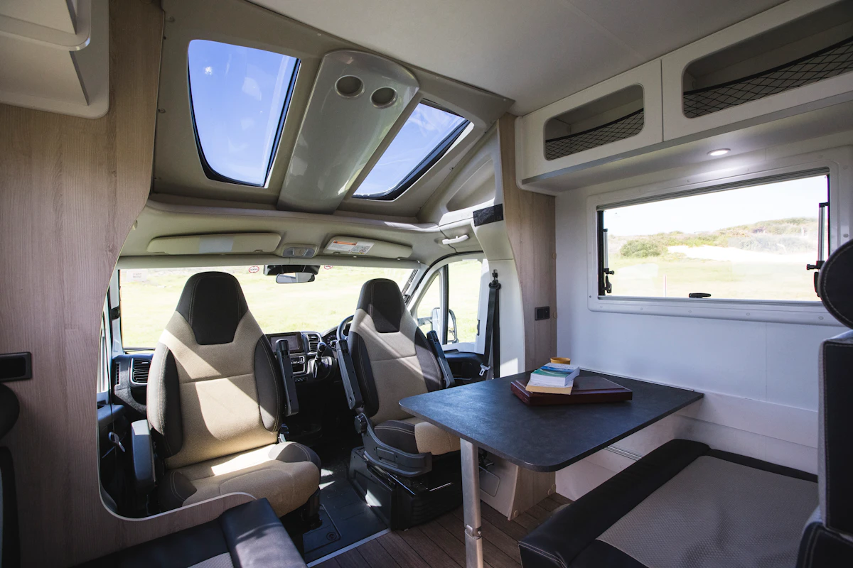 Modern campervan interior with two swivel seats, a dining table, skylights, and a window with scenic outdoor view.