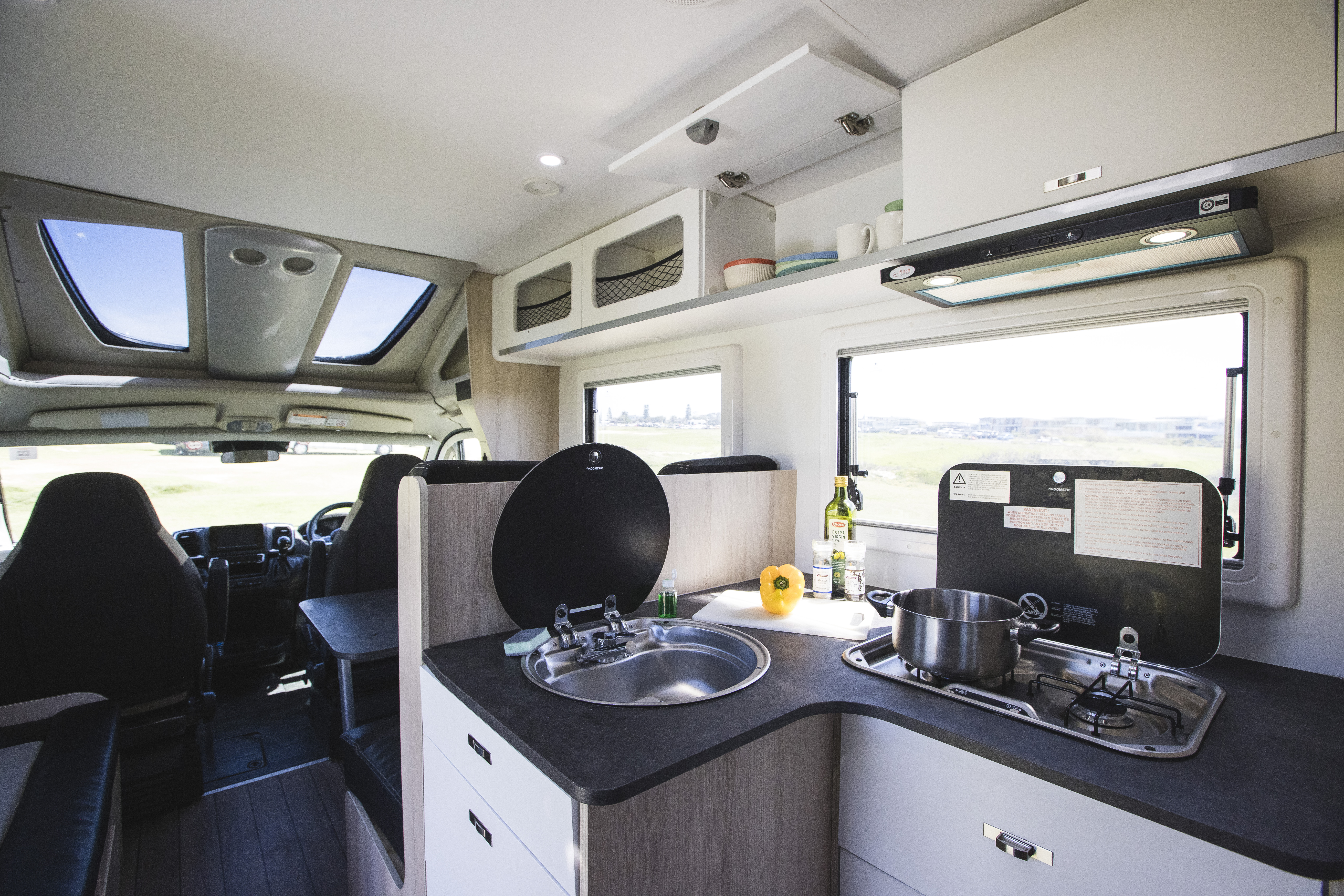 Modern campervan interior featuring a compact kitchen with sink, stove, pot, and open window overlooking scenic landscape.