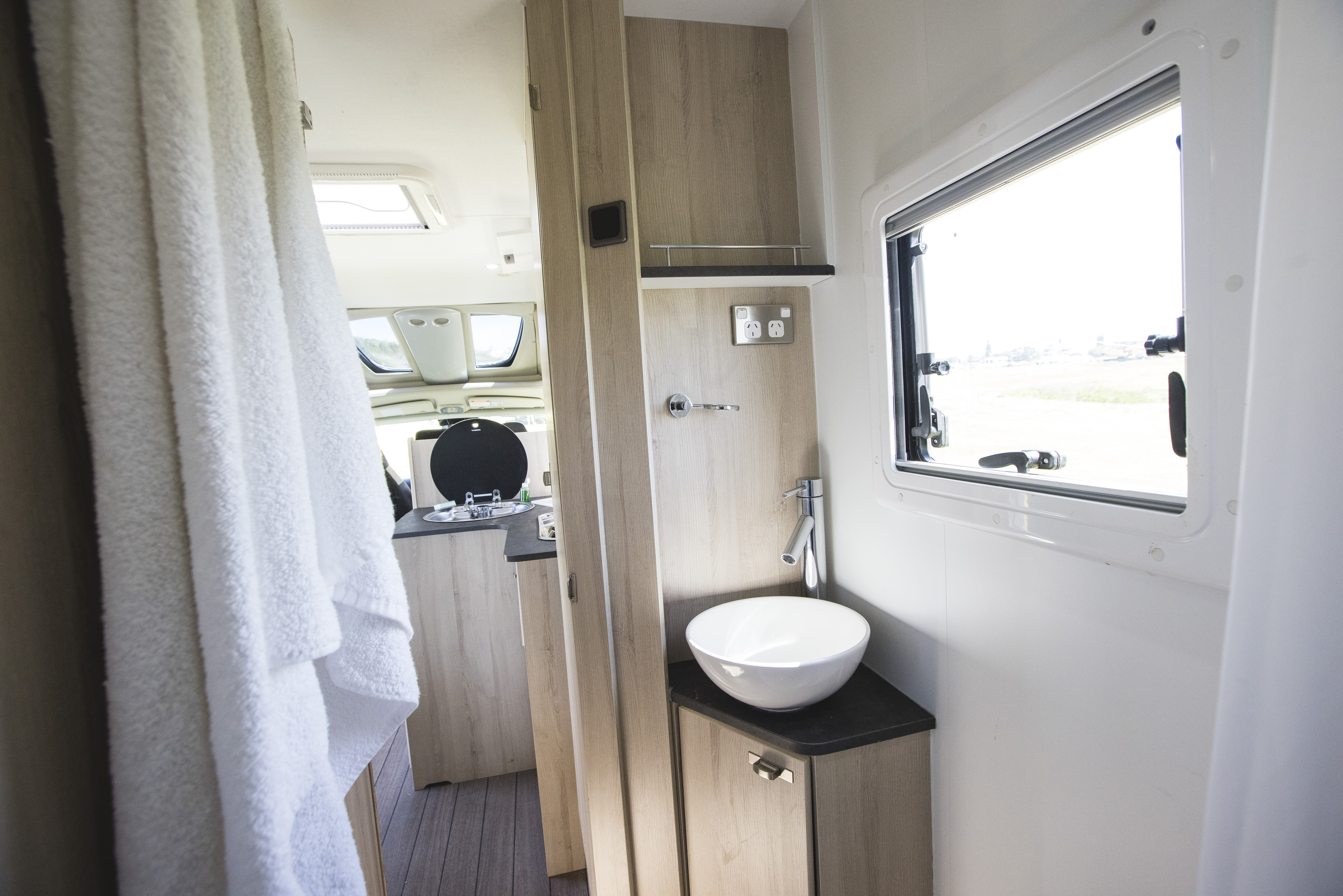 Modern campervan bathroom with white sink, wooden cabinets, towels, and window, leading to kitchen and dining area.