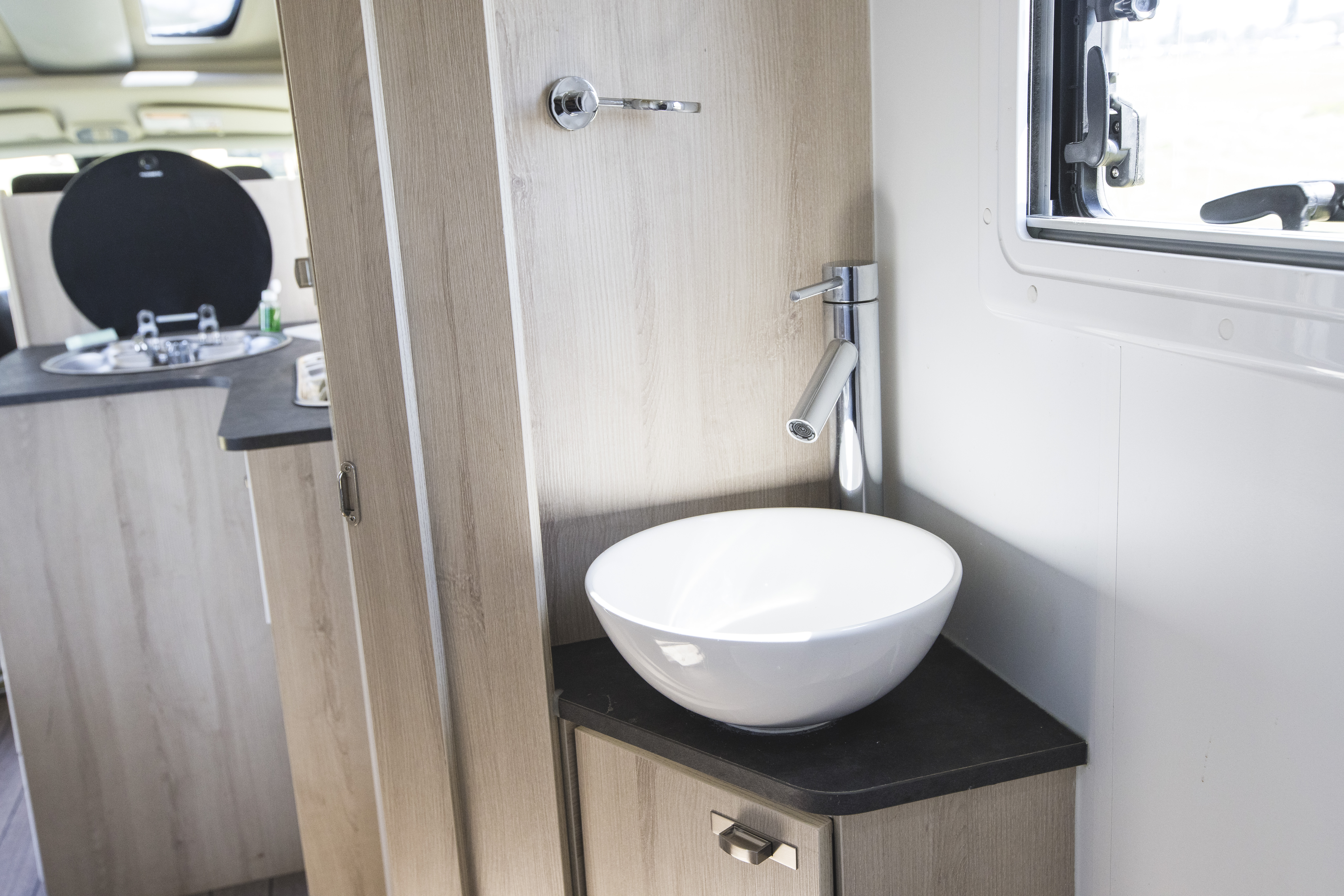 Modern campervan bathroom with a white bowl sink, chrome tap, wooden cabinets, and window with natural light.