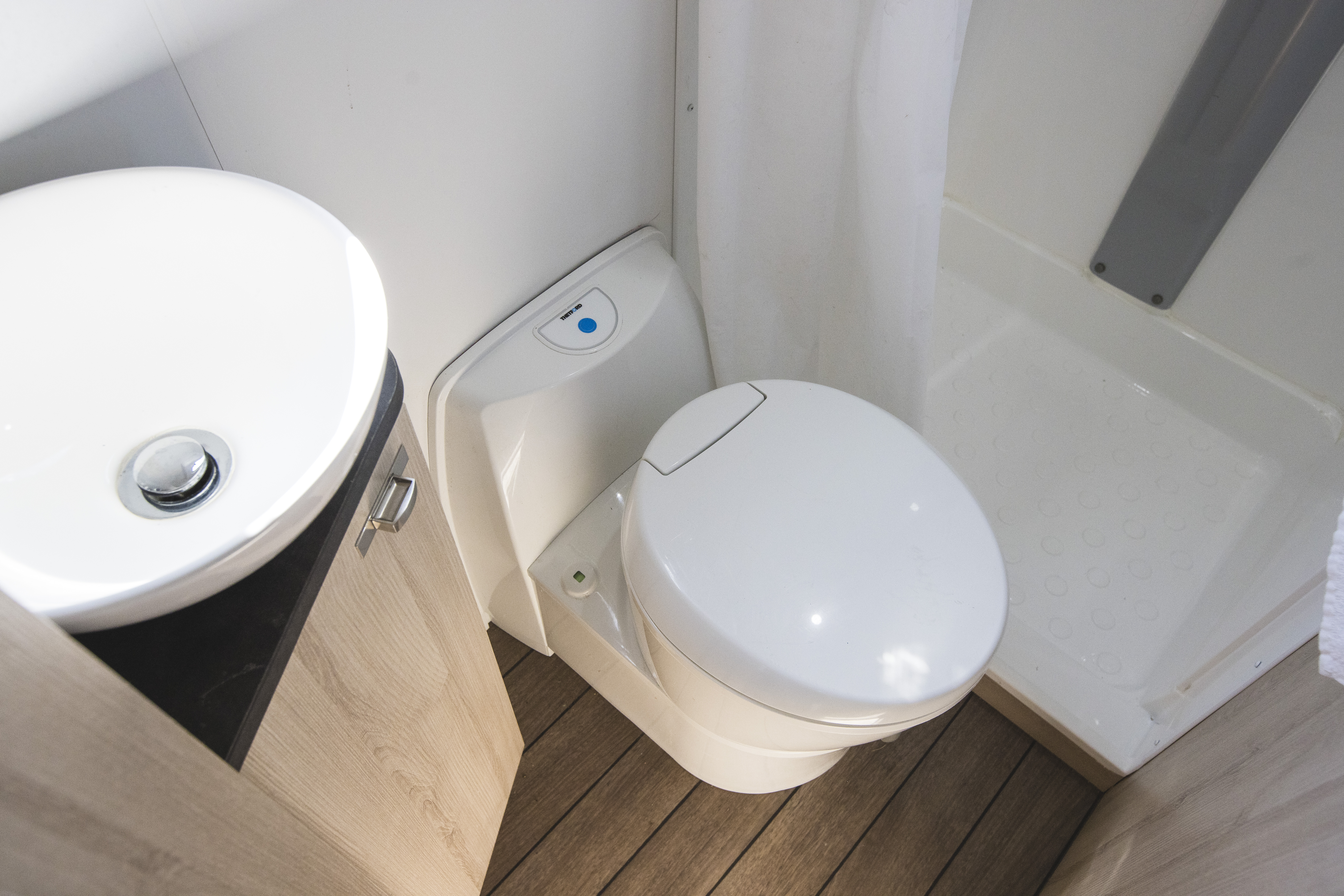 Compact campervan bathroom with a white toilet, small round sink, wooden floor, and shower area.