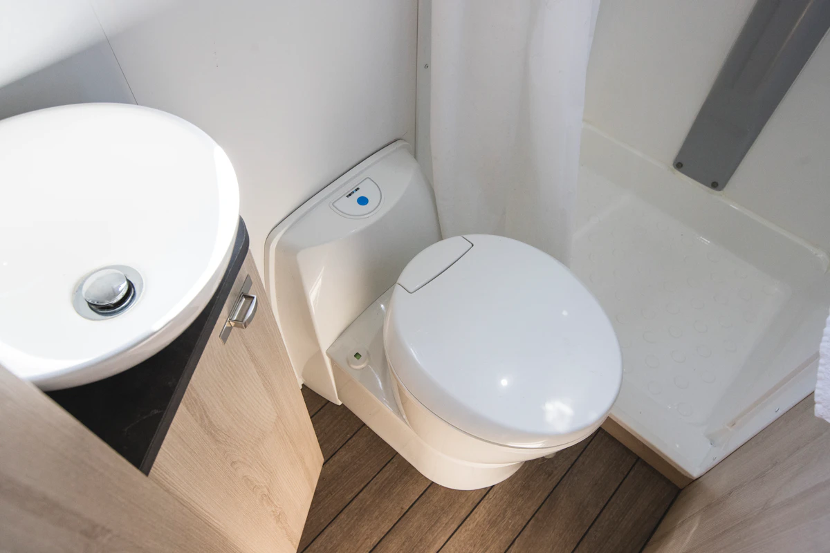 Compact campervan bathroom with a white toilet, small round sink, wooden floor, and shower area.