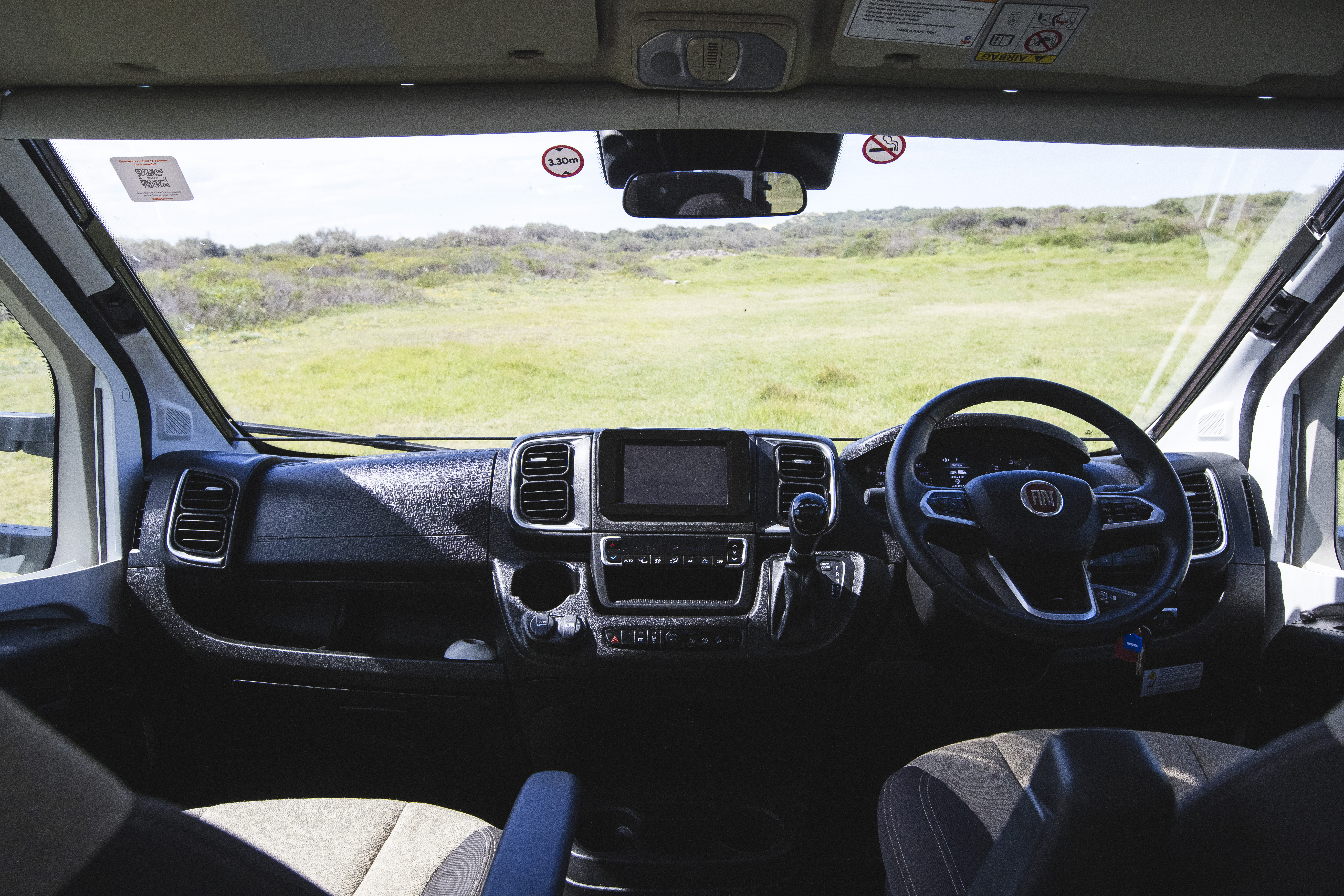 View from inside a campervan showing the dashboard, steering wheel, and grassy landscape through the windshield.