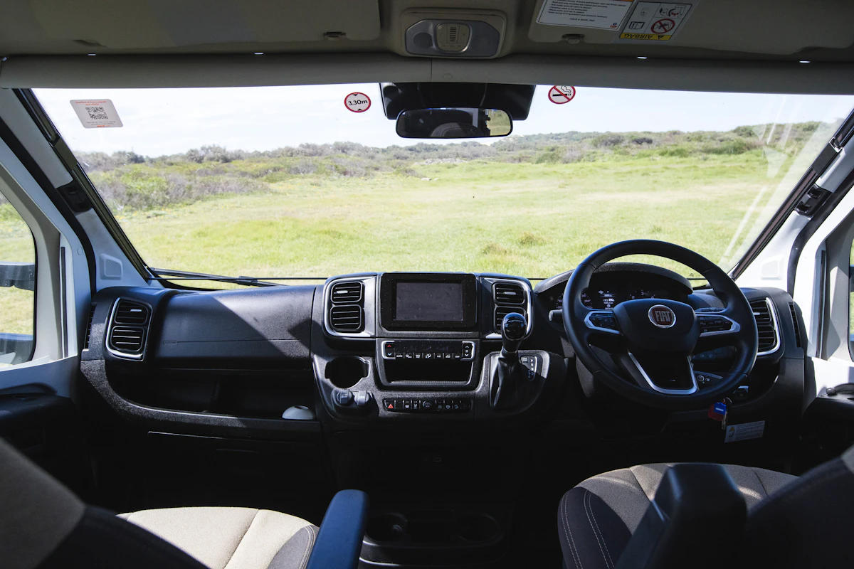 View from inside a campervan showing the dashboard, steering wheel, and grassy landscape through the windshield.