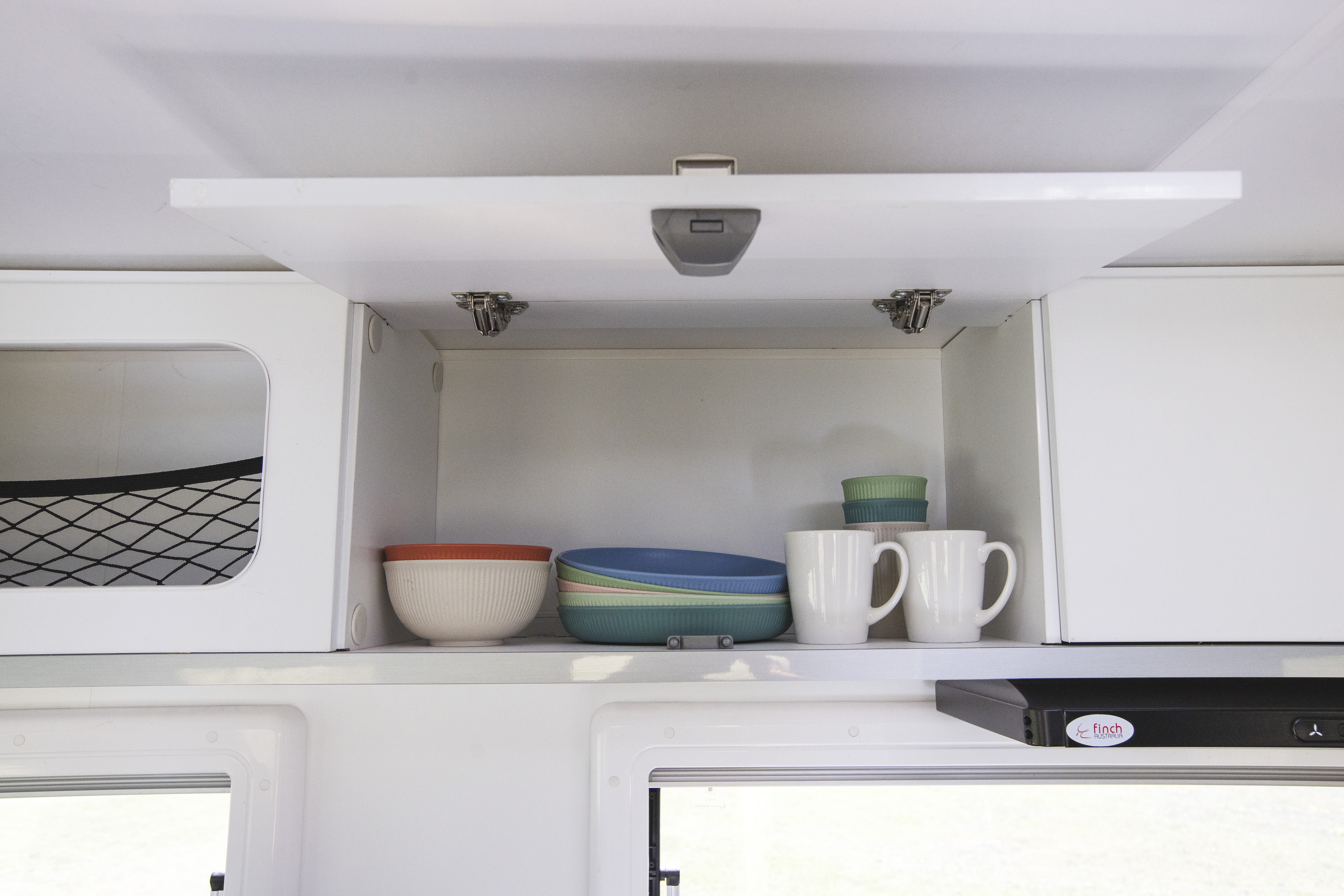 Open campervan cupboard showing stacked bowls, plates, and two white mugs on a white shelf.