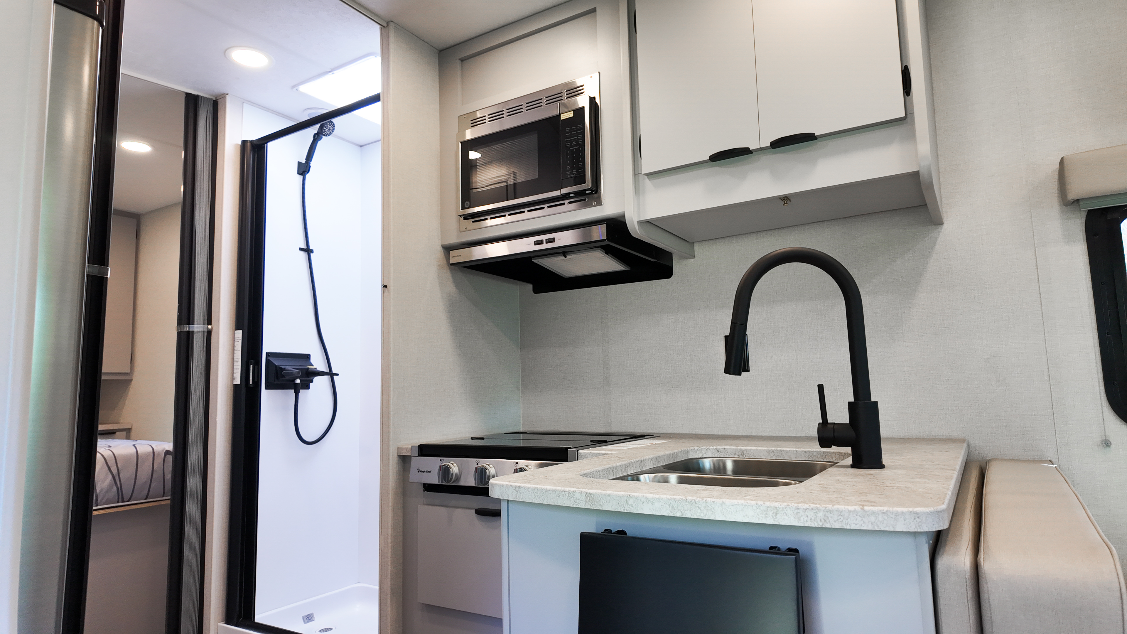 Modern camper van interior with kitchenette, sink, stove, microwave, overhead cabinets, and shower area visible.