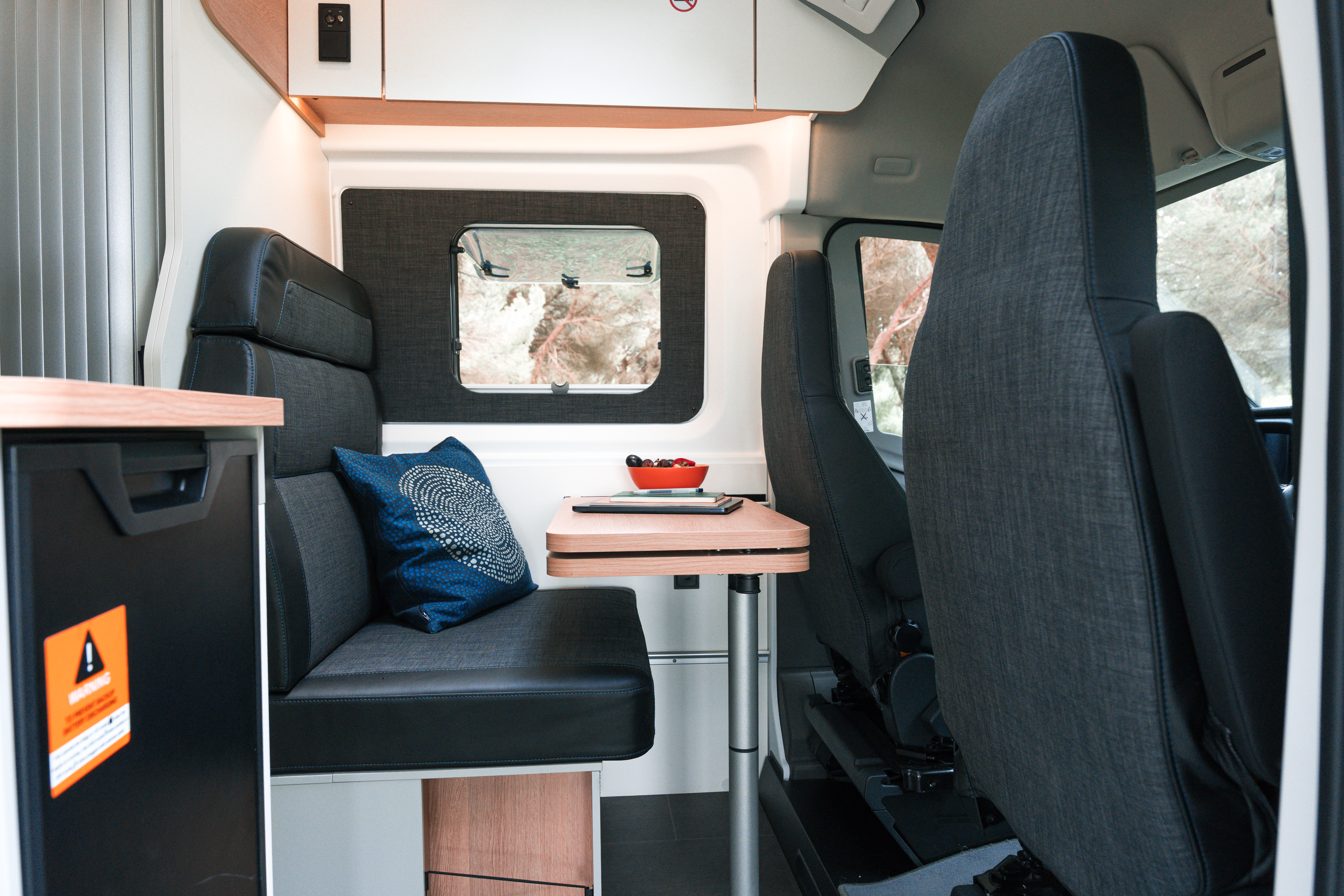 Intérieur d’un van aménagé avec banquette, table, coussin décoratif et fenêtre, idéal pour camping et voyages.
