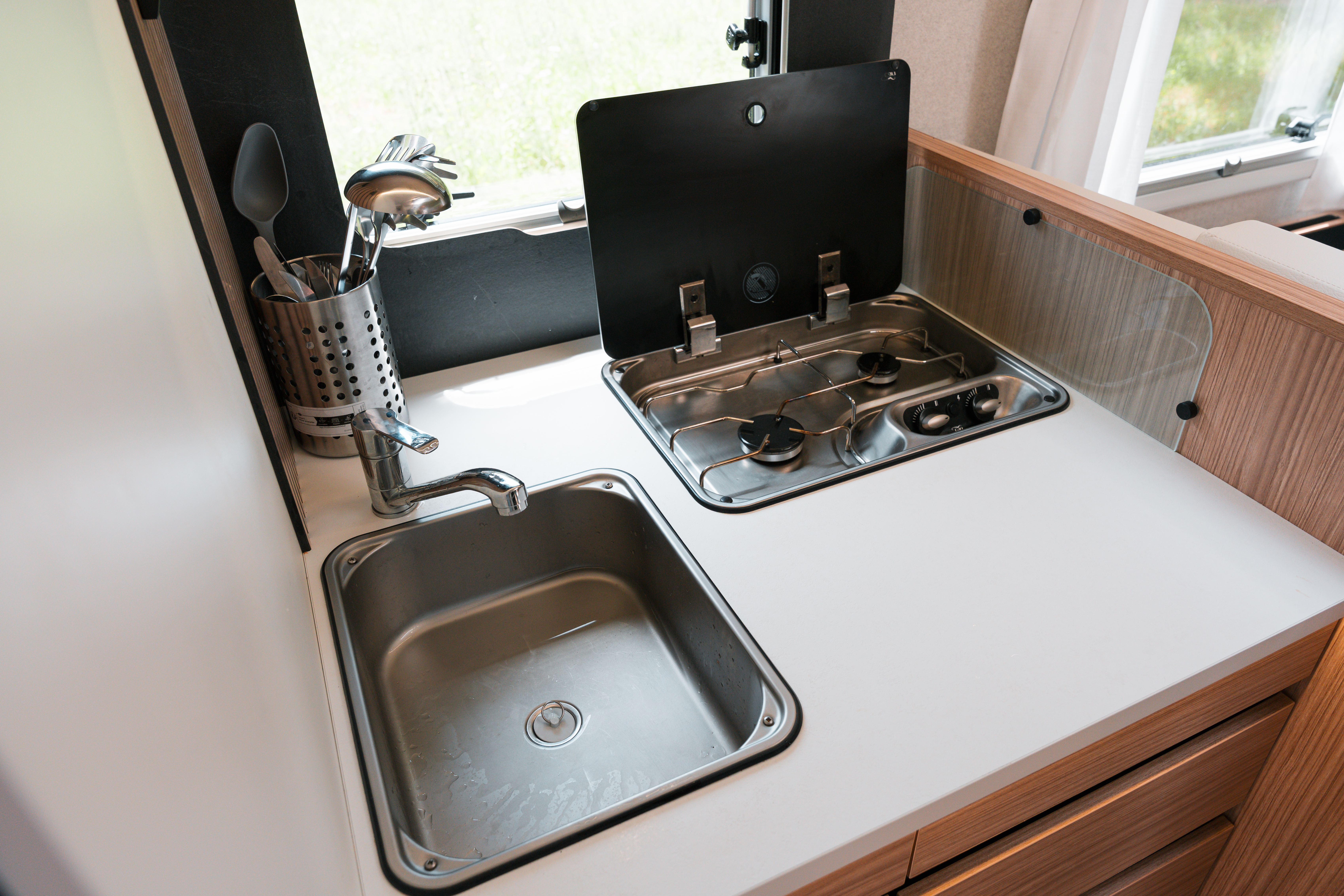 Compact camper van kitchen with stainless steel sink, gas stove, faucet, and utensil holder on a white countertop.