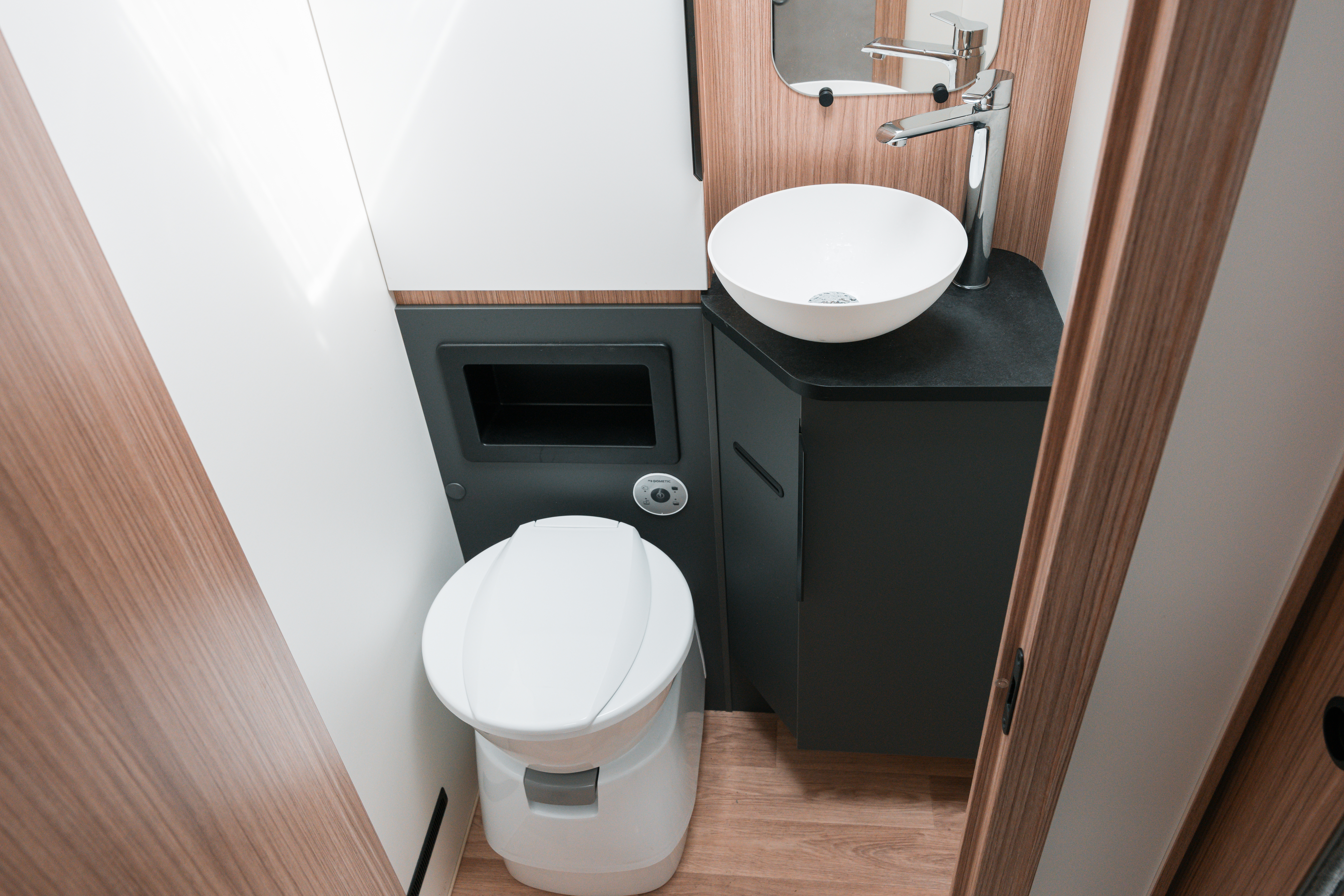 Modern campervan bathroom with a toilet, sink, mirror, wood paneling, and compact black counter.