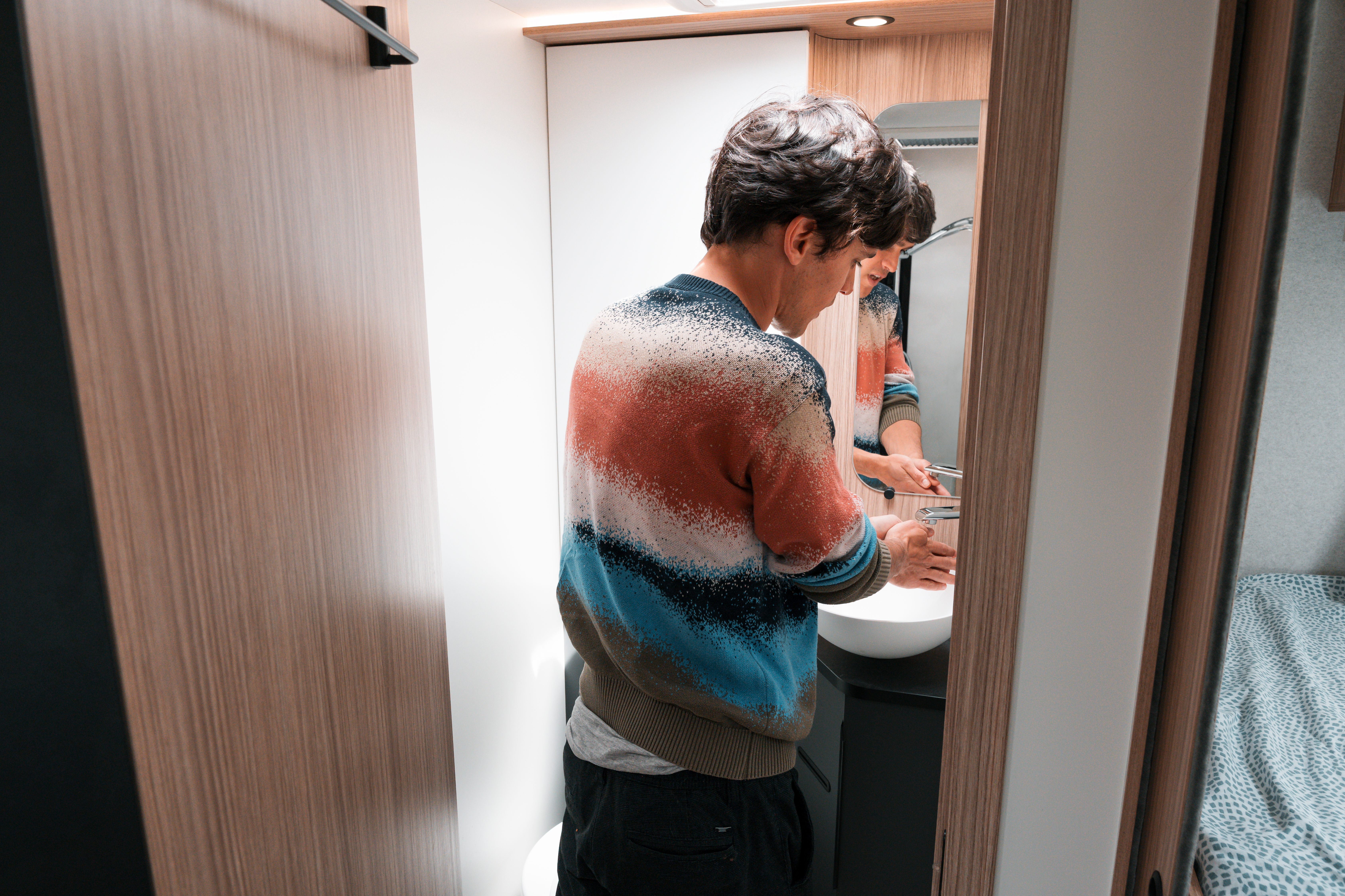 Person washing hands at a compact campervan bathroom sink with a mirror and wooden paneling.
