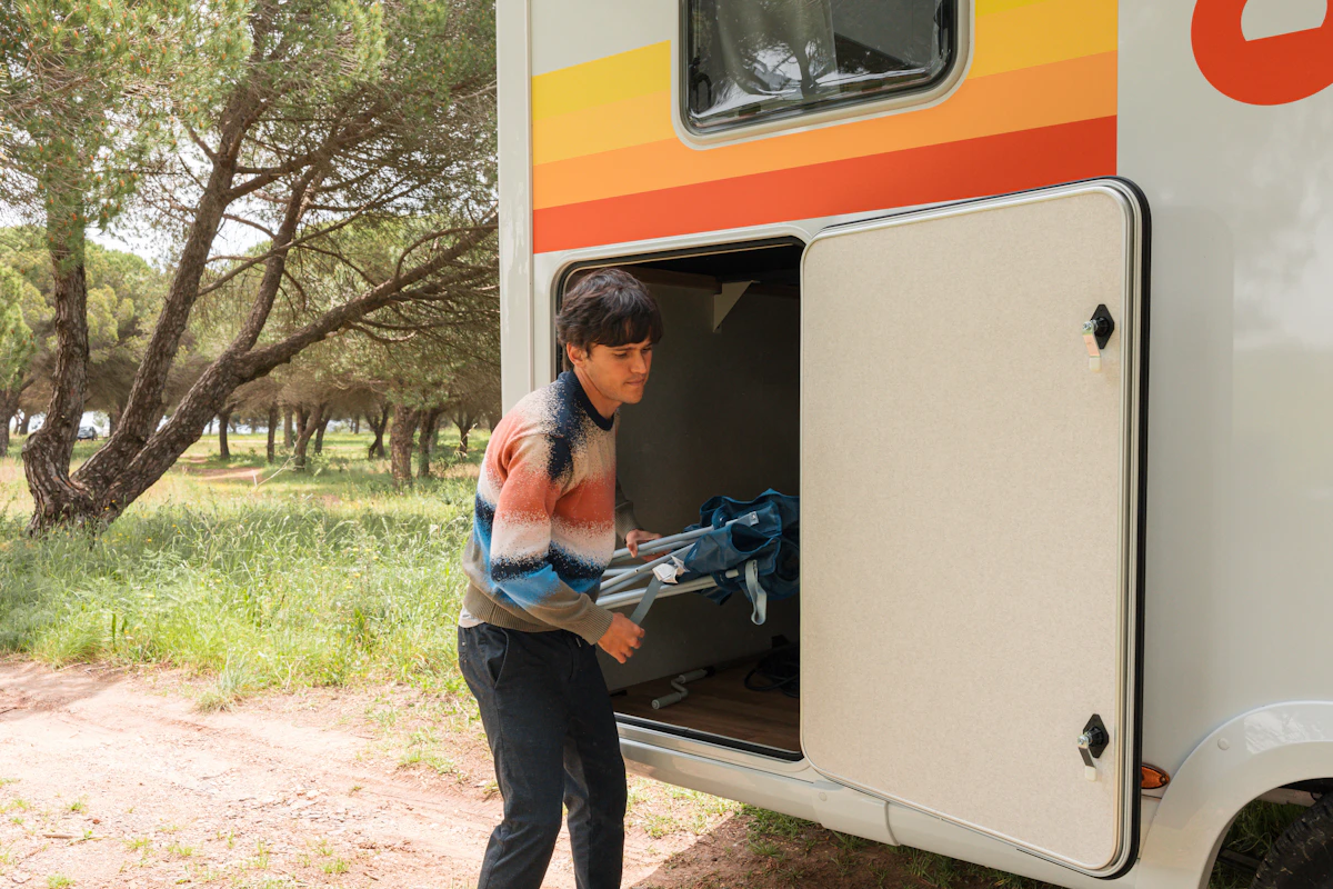 Person loading camping chairs into the storage compartment of a camper van parked near trees in a grassy area.