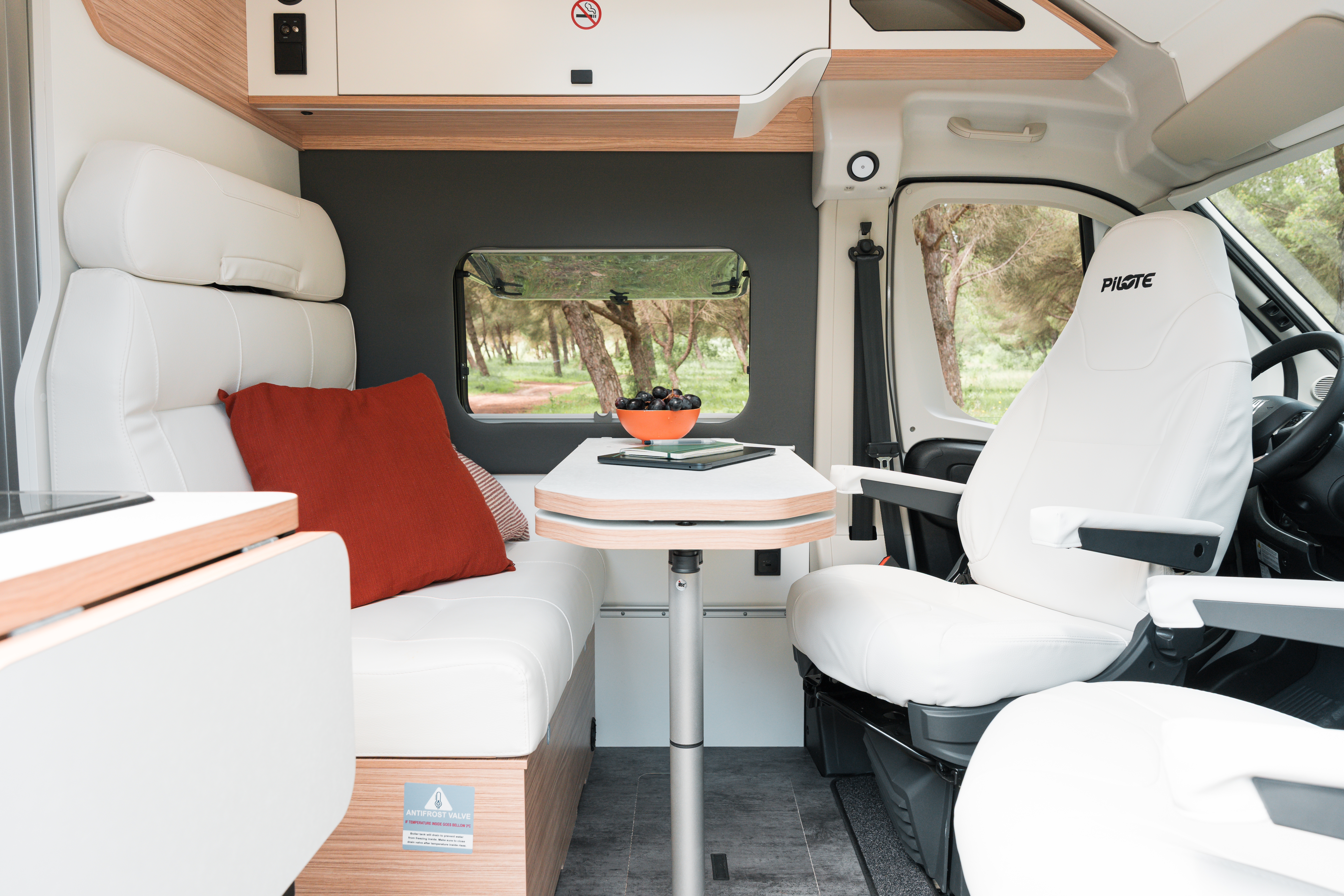 Modern campervan interior with white seats, table, red pillow, and window view of trees in the background.