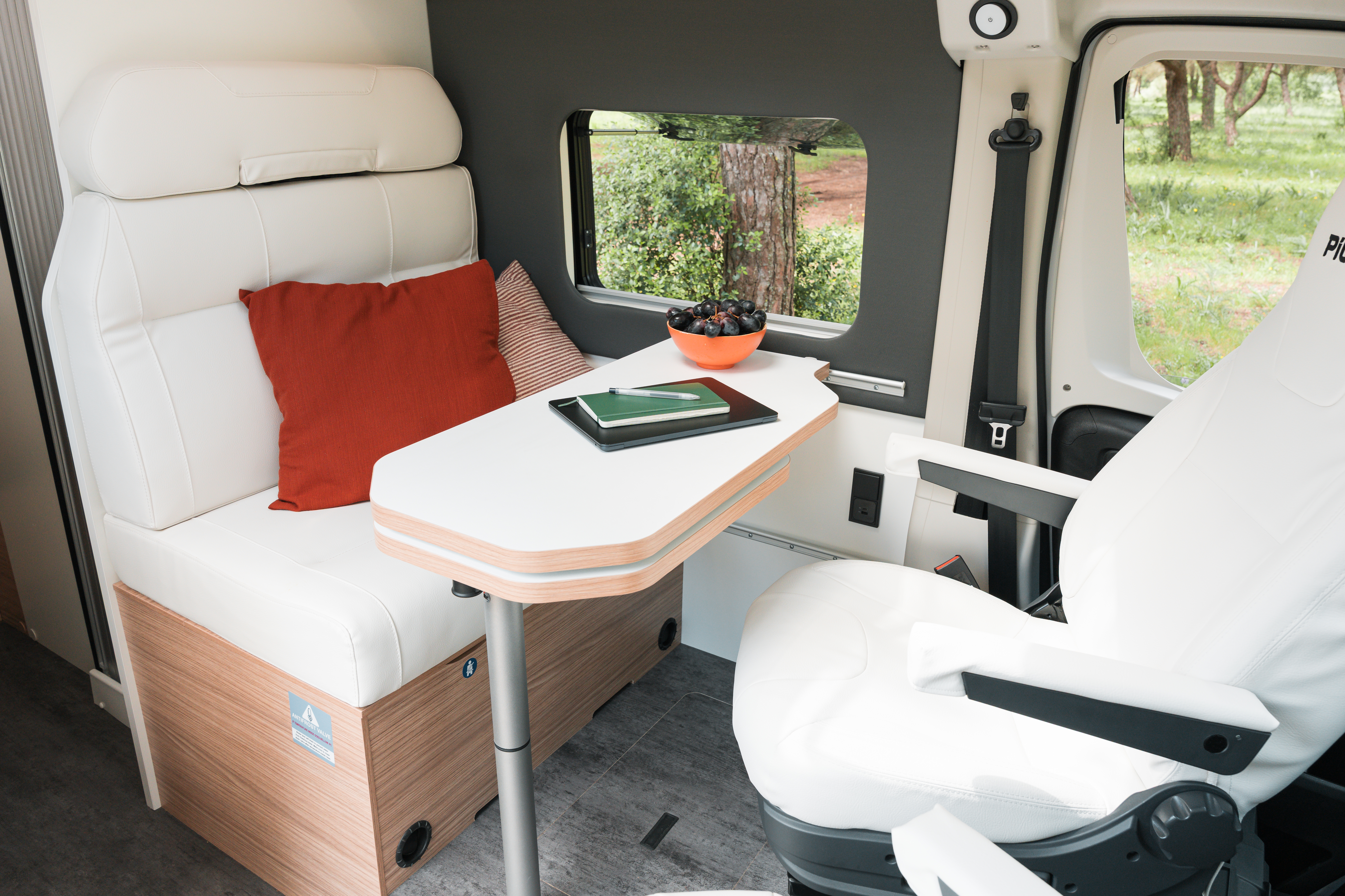 Interior of a campervan with a table, two white seats, an orange pillow, and a bowl of berries.