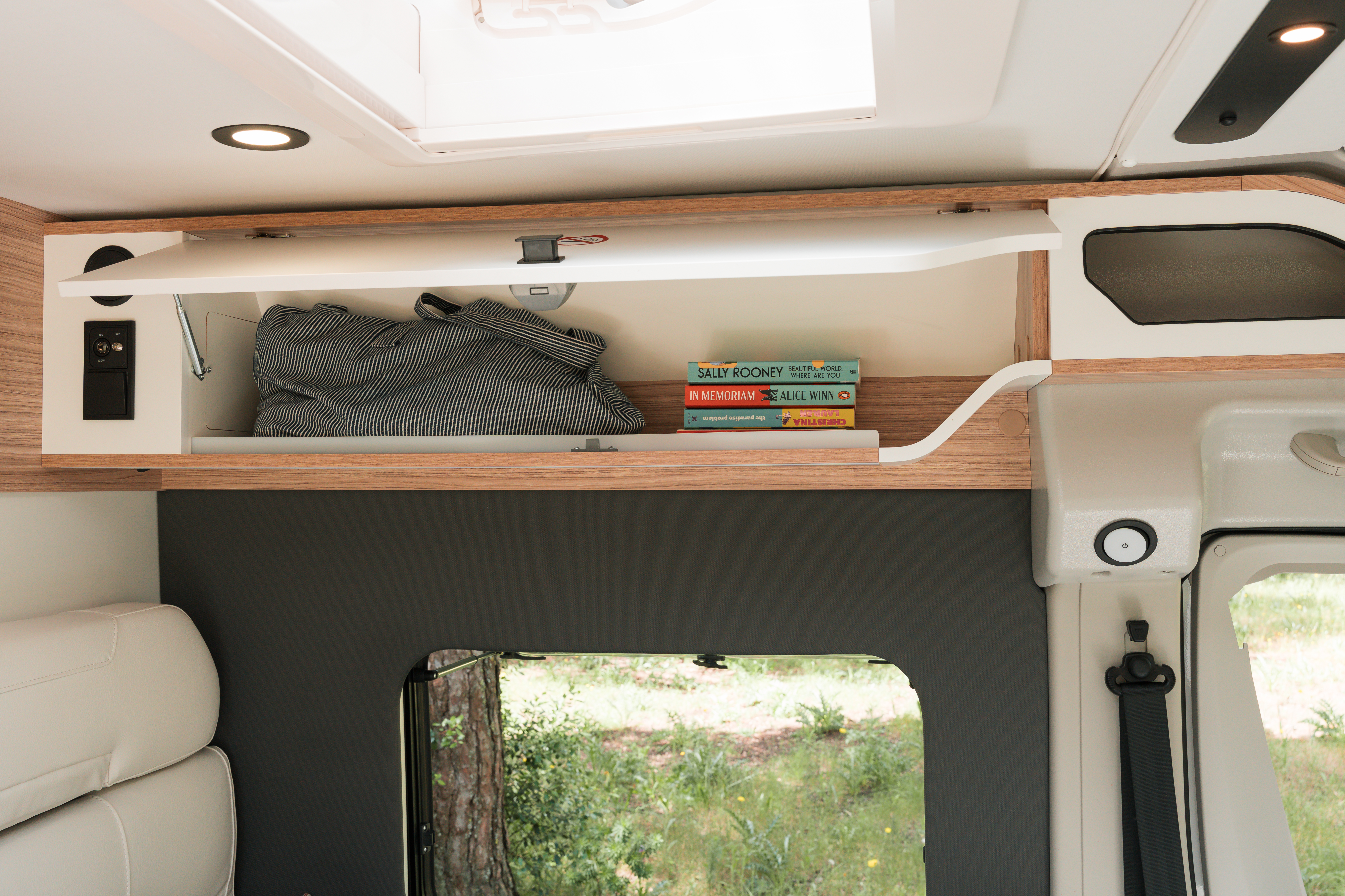 Open overhead storage compartment in a camper van with folded clothes and several books neatly placed inside.