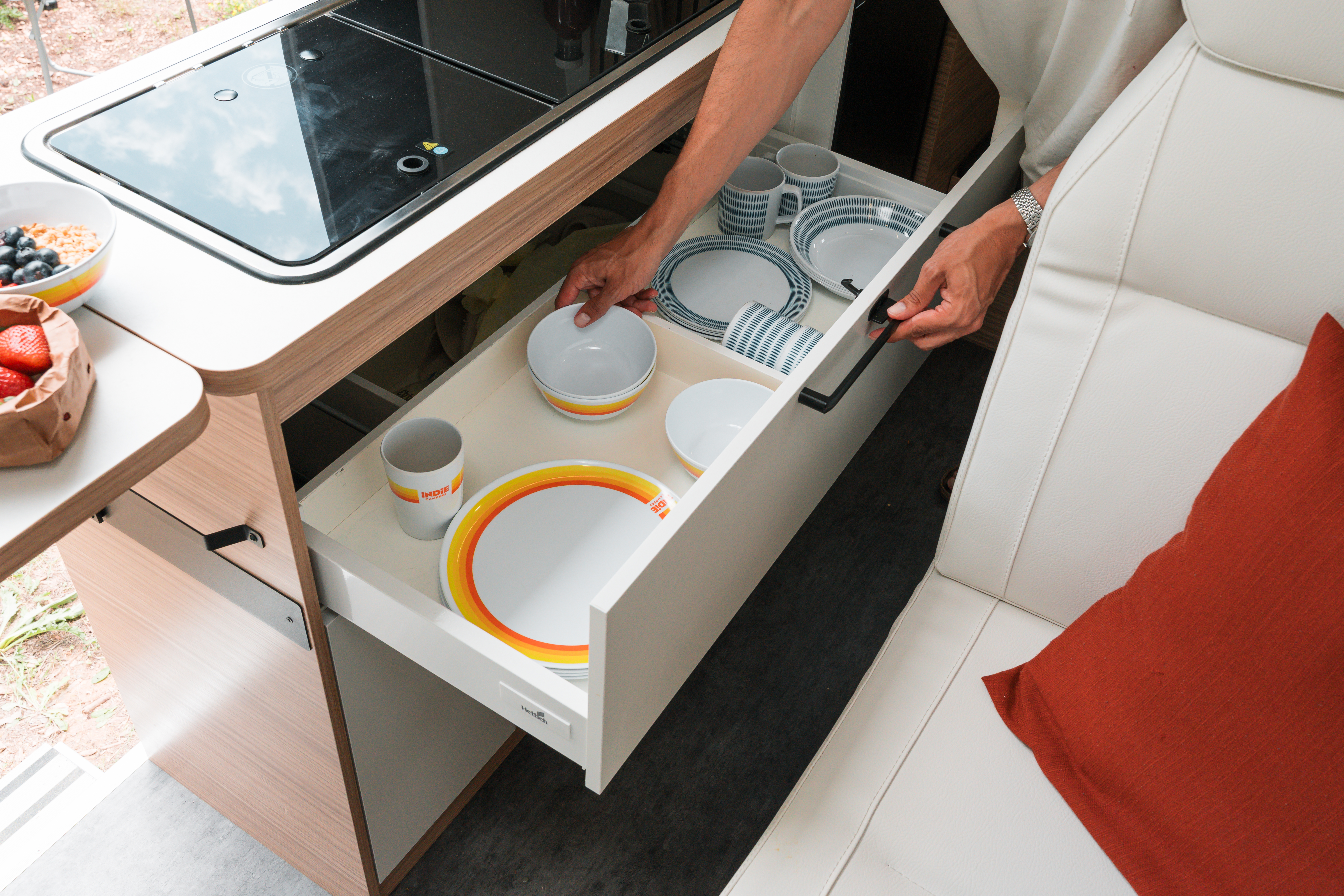 Person opening a kitchen drawer filled with plates and cups inside a campervan next to a white seat.
