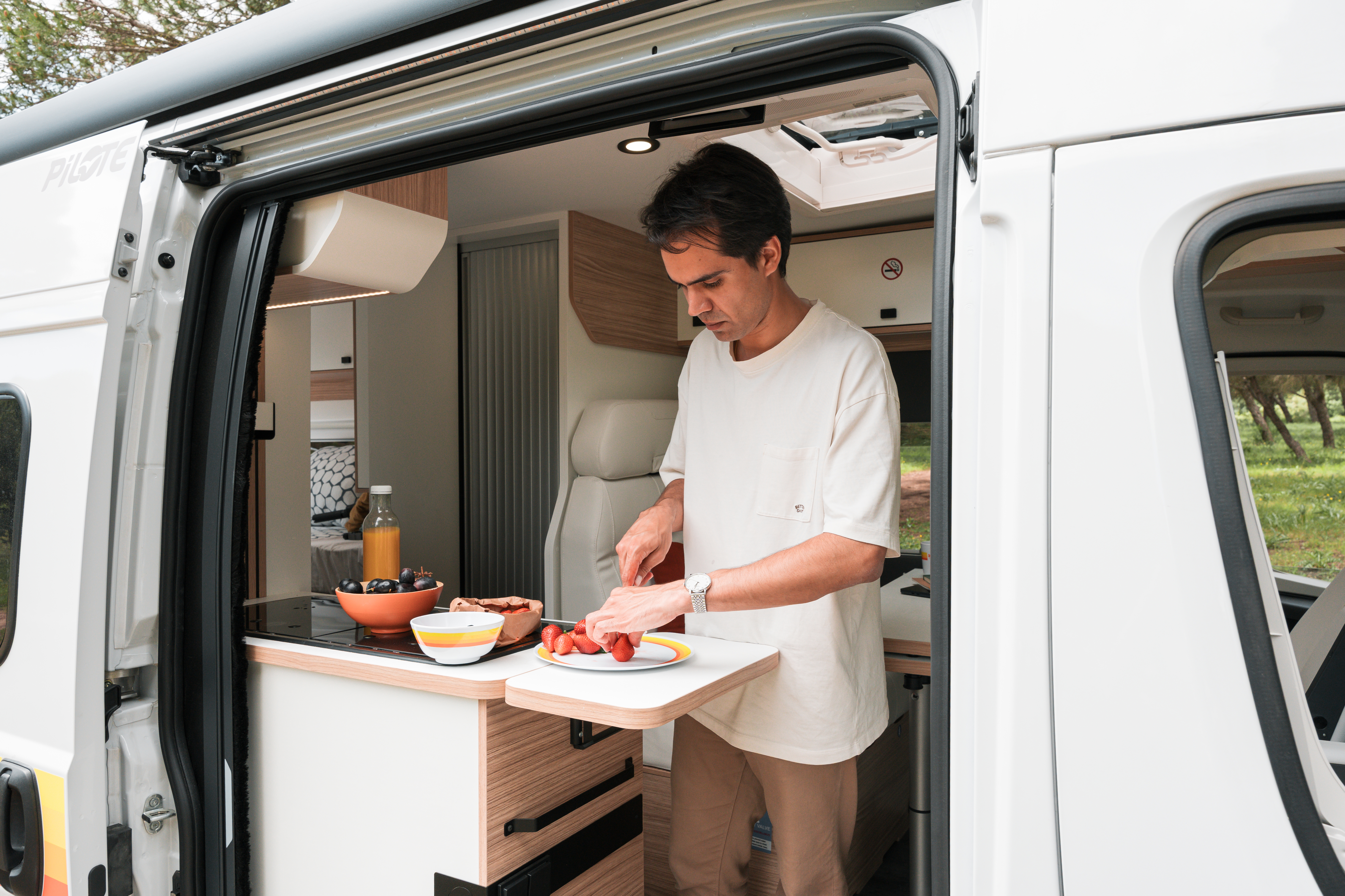 Man slicing fruit in the kitchen area of a camper van with ingredients and juice on the counter.