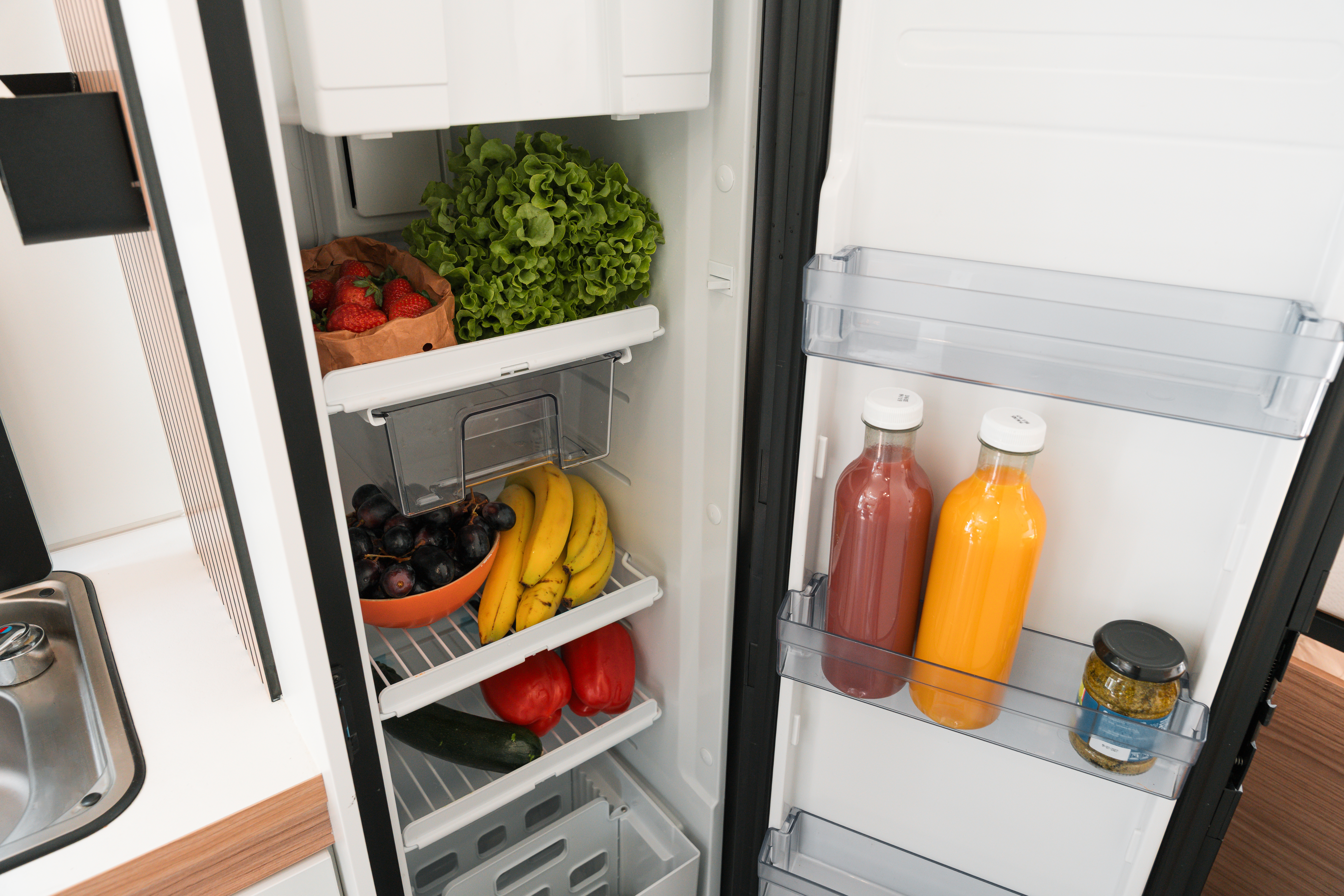 Open campervan fridge filled with fresh fruits, vegetables, juice bottles, and a jar, showcasing campervan kitchen amenities.