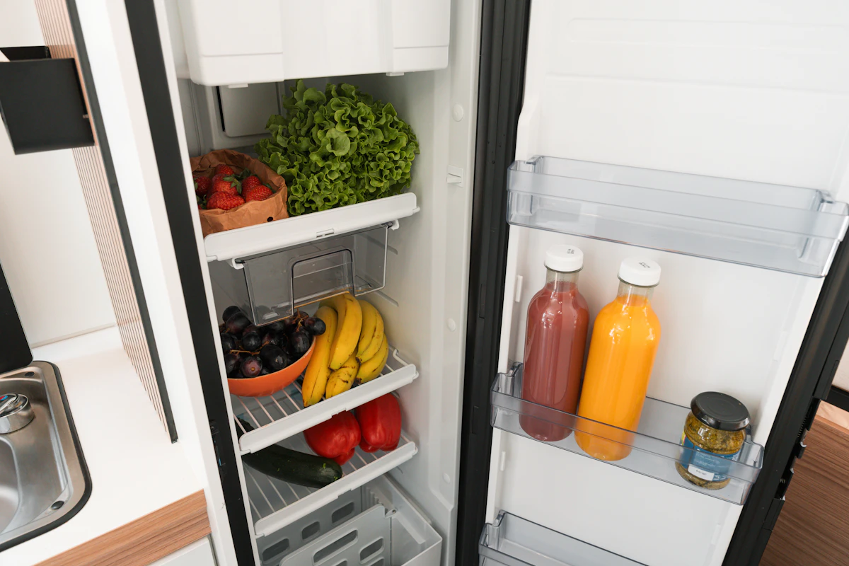 Open campervan fridge filled with fresh fruits, vegetables, juice bottles, and a jar, showcasing campervan kitchen amenities.