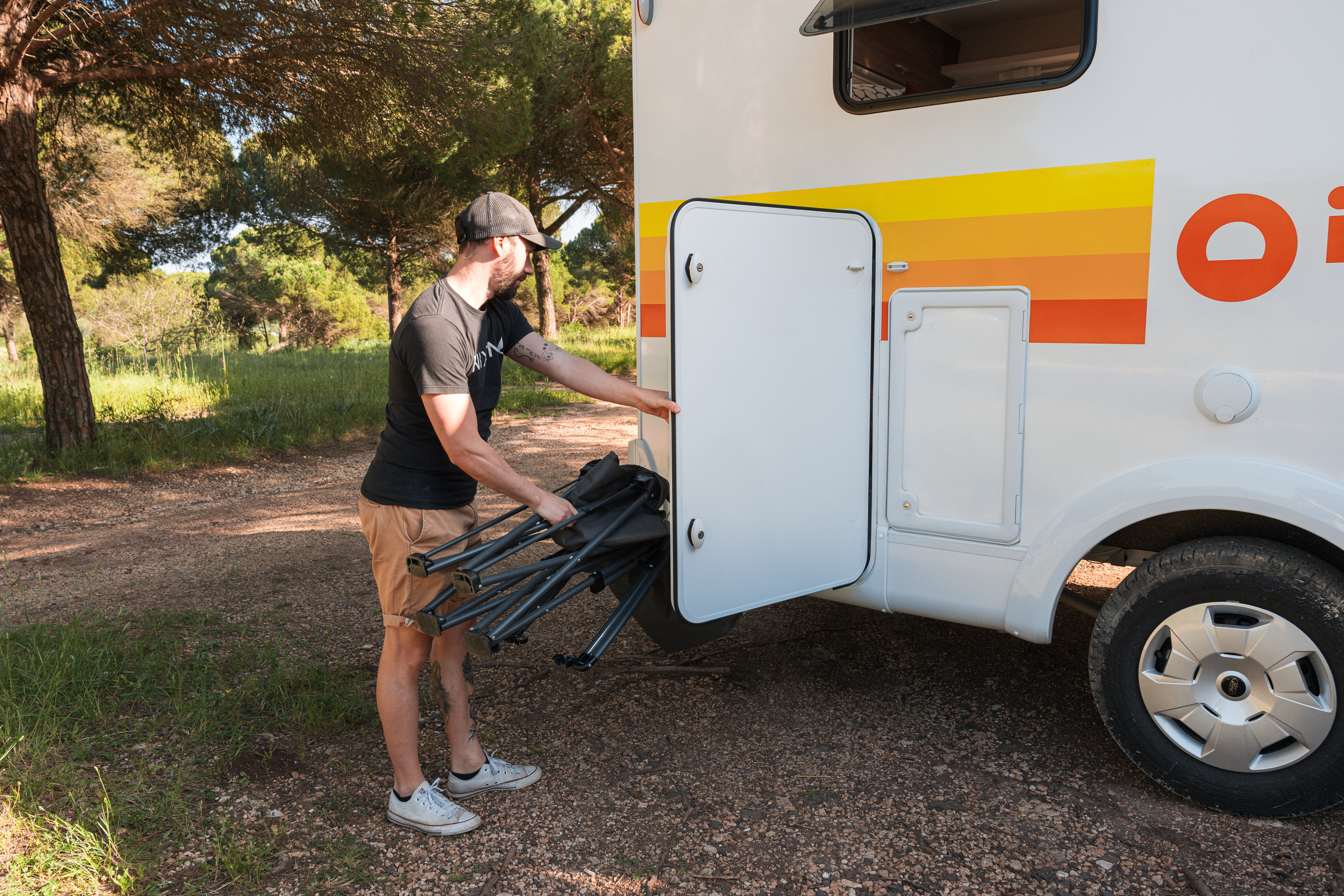 Person loading folding chairs into a storage compartment on the side of a campervan parked outdoors.
