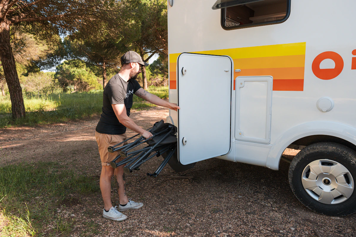 Person loading folding chairs into a storage compartment on the side of a campervan parked outdoors.
