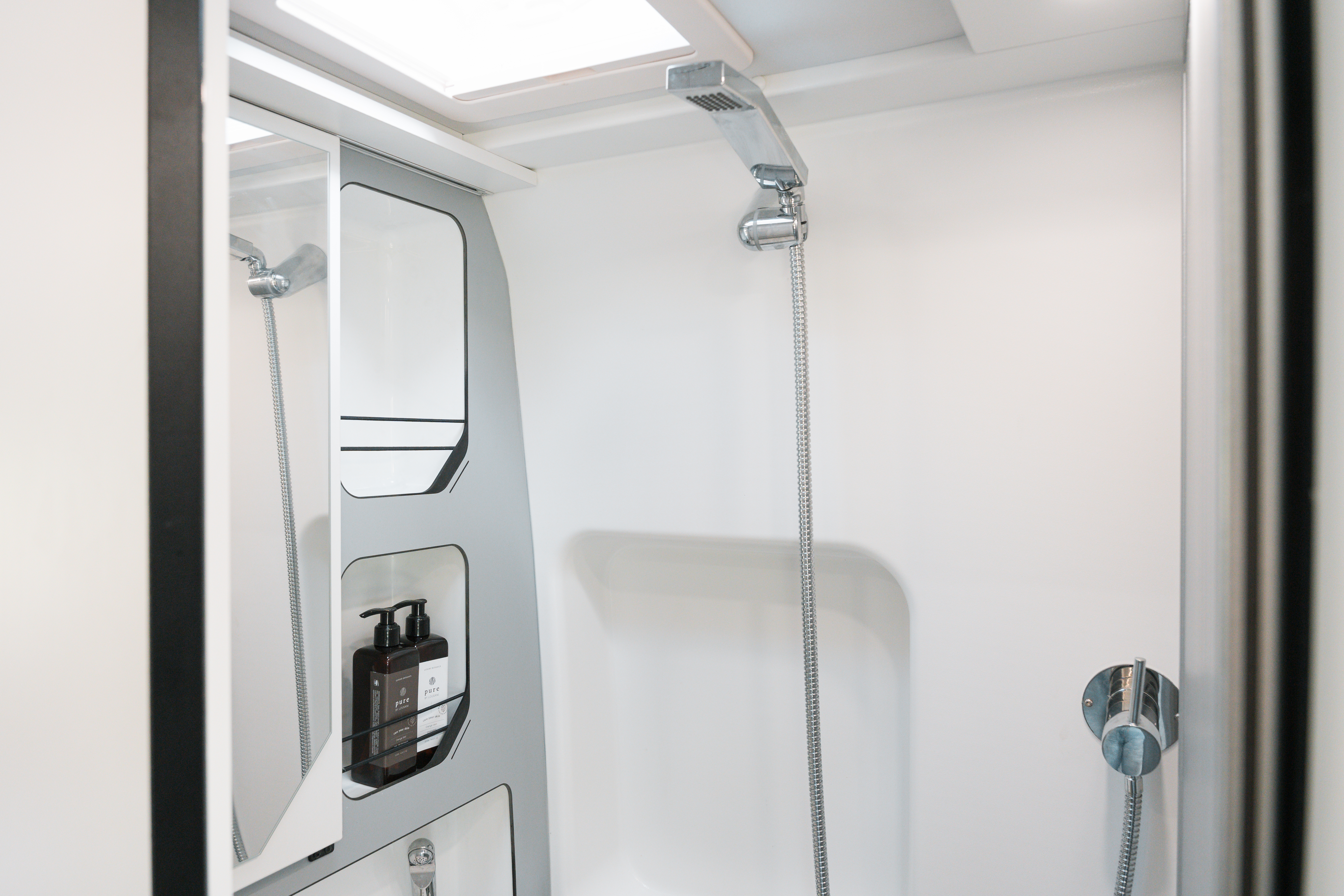 Modern campervan shower with overhead skylight, wall mirror, mounted showerhead, and soap dispensers on a white interior.