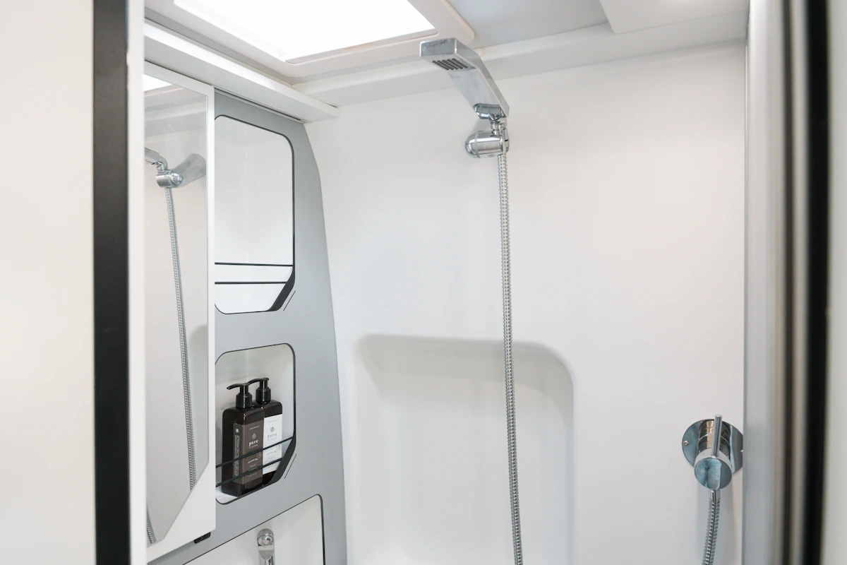 Modern campervan shower with overhead skylight, wall mirror, mounted showerhead, and soap dispensers on a white interior.