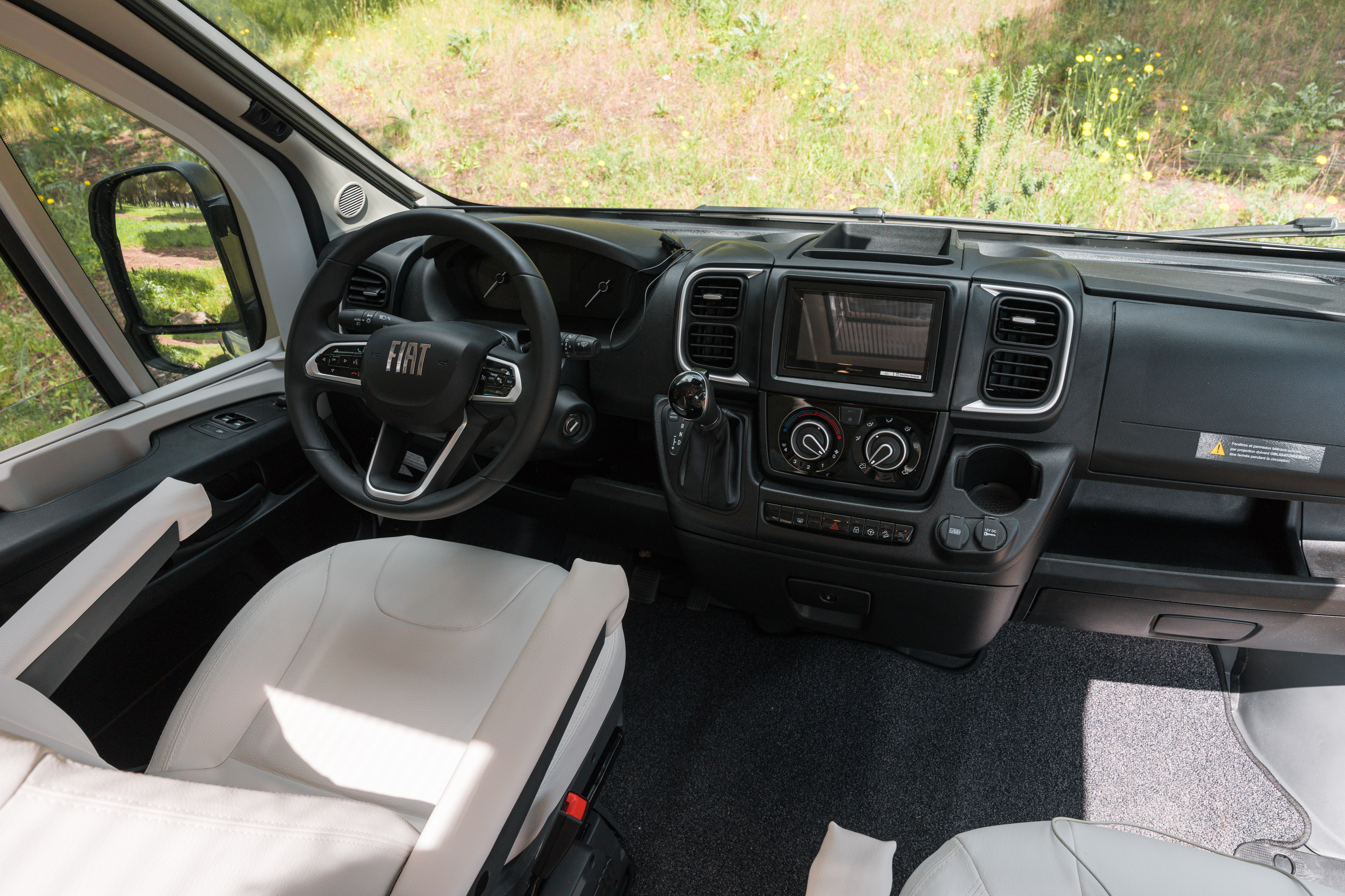 Interior view of a Fiat camper van cabin with two seats, steering wheel, dashboard, and forest background.