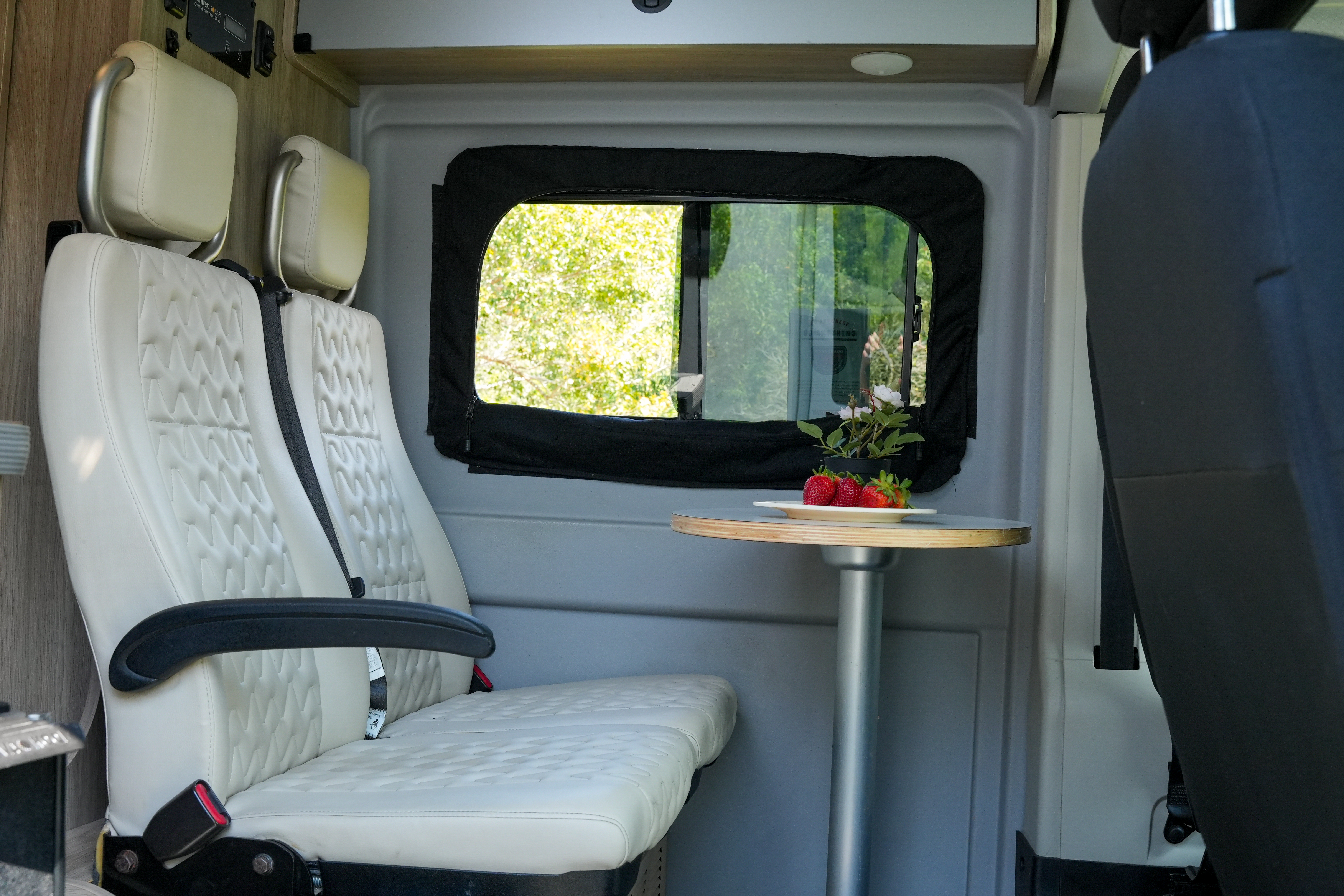 Interior of a camper van with two cream seats, a small table, and a window showing outside greenery.