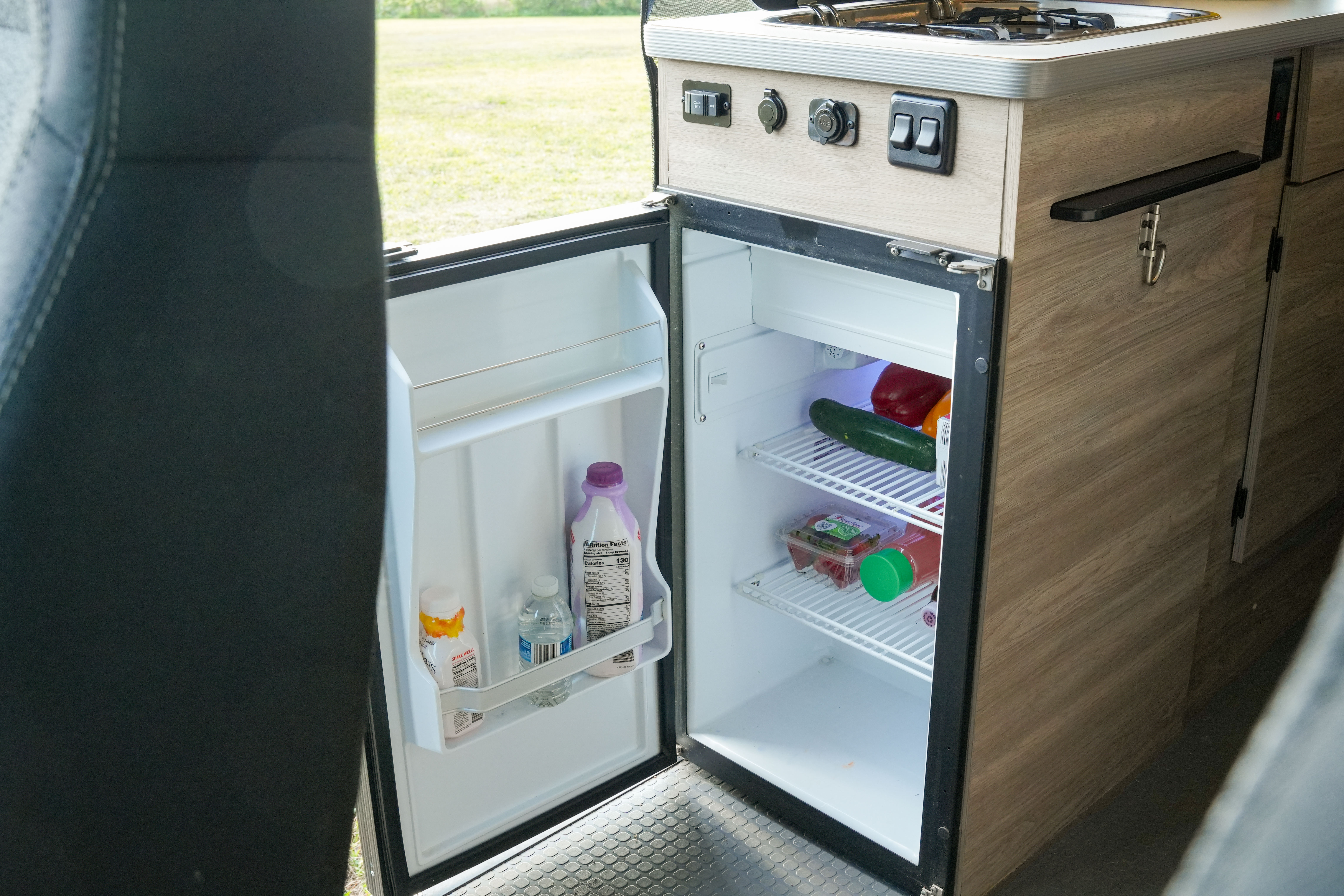 Small campervan fridge open, showing drinks, vegetables and groceries inside, next to a compact kitchenette.