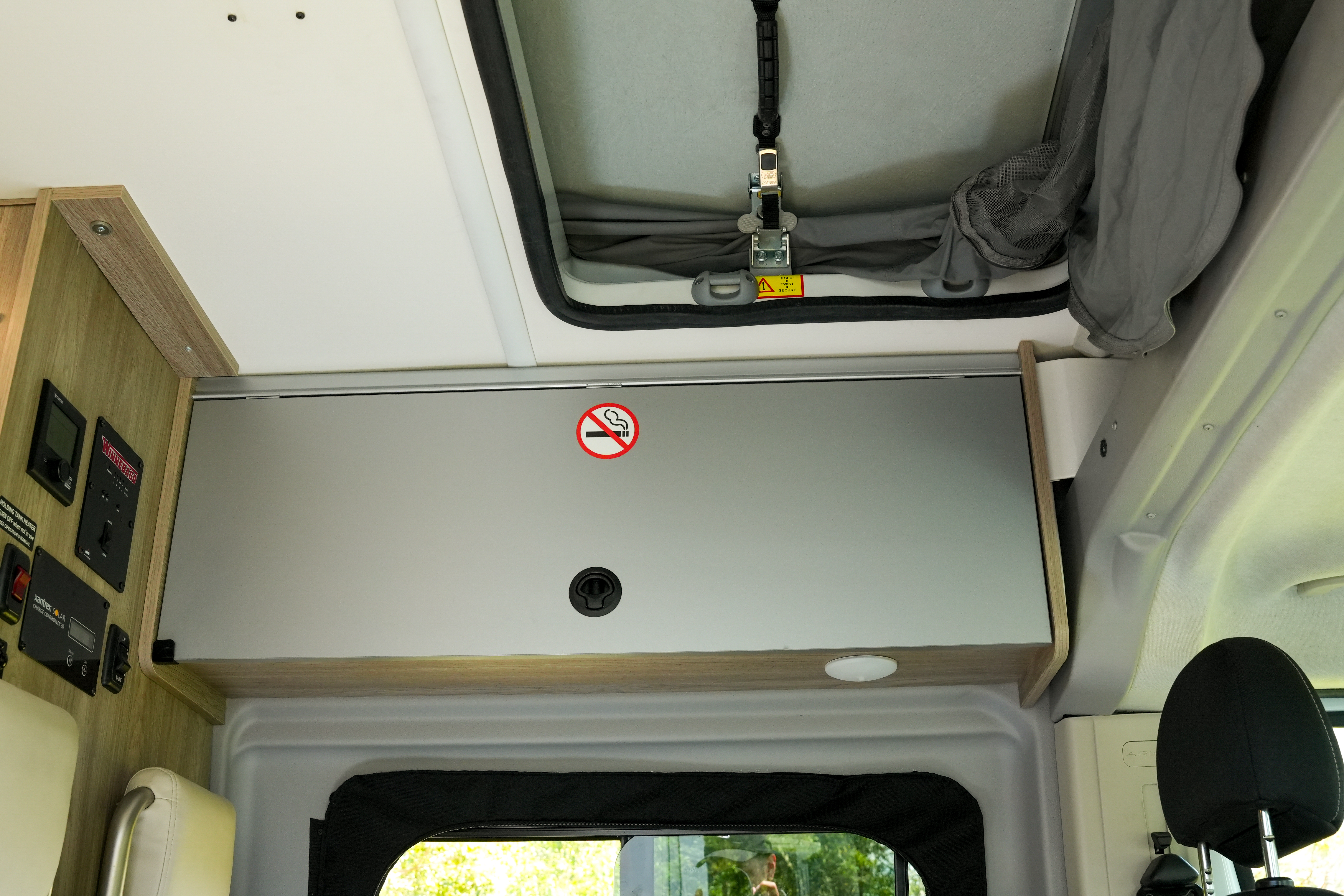 Interior of a campervan showing an overhead storage cabinet, control panels, and a no smoking sign near a roof hatch.