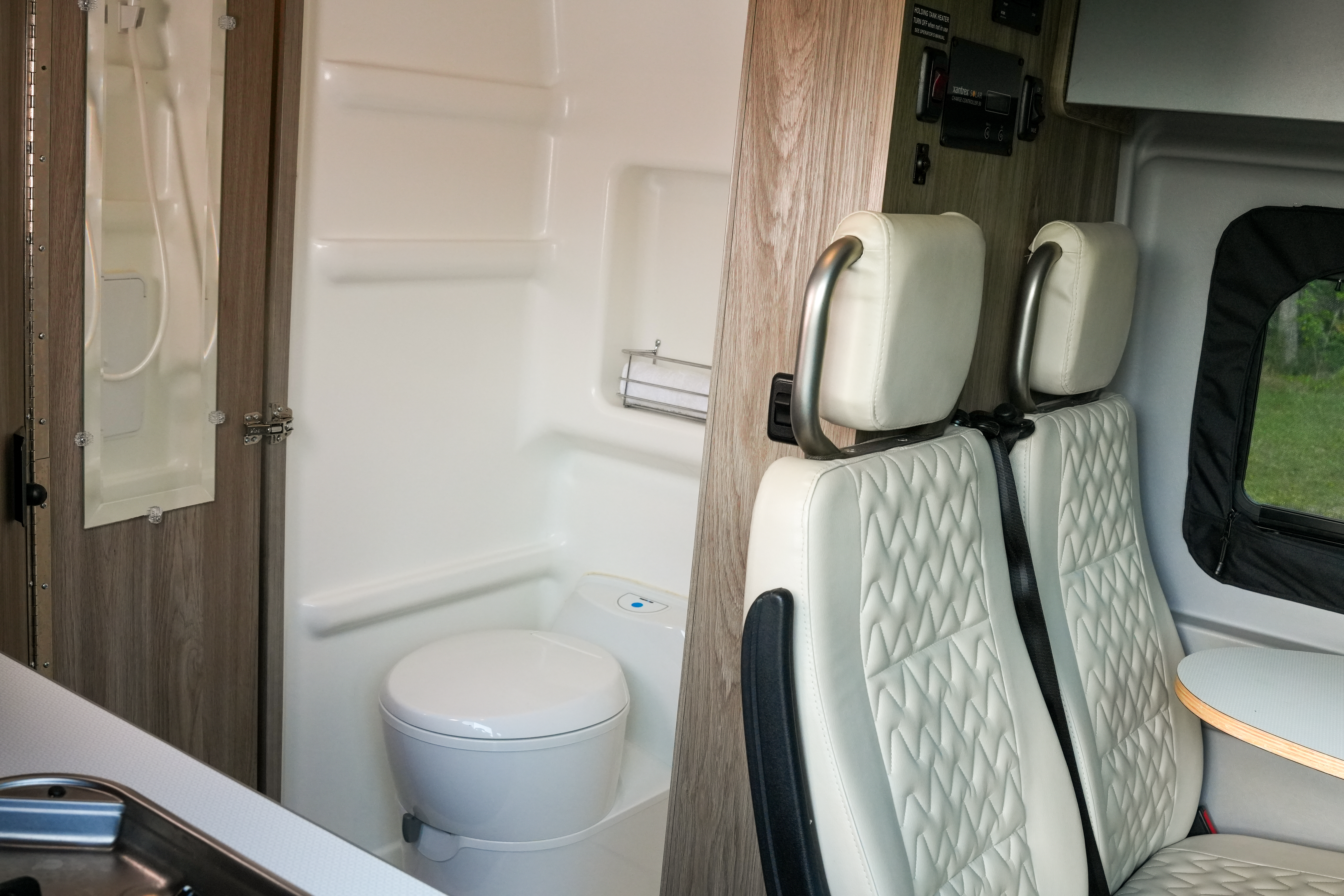 Campervan interior with a compact bathroom, toilet, two swivel seats, and a small window with outdoor view.