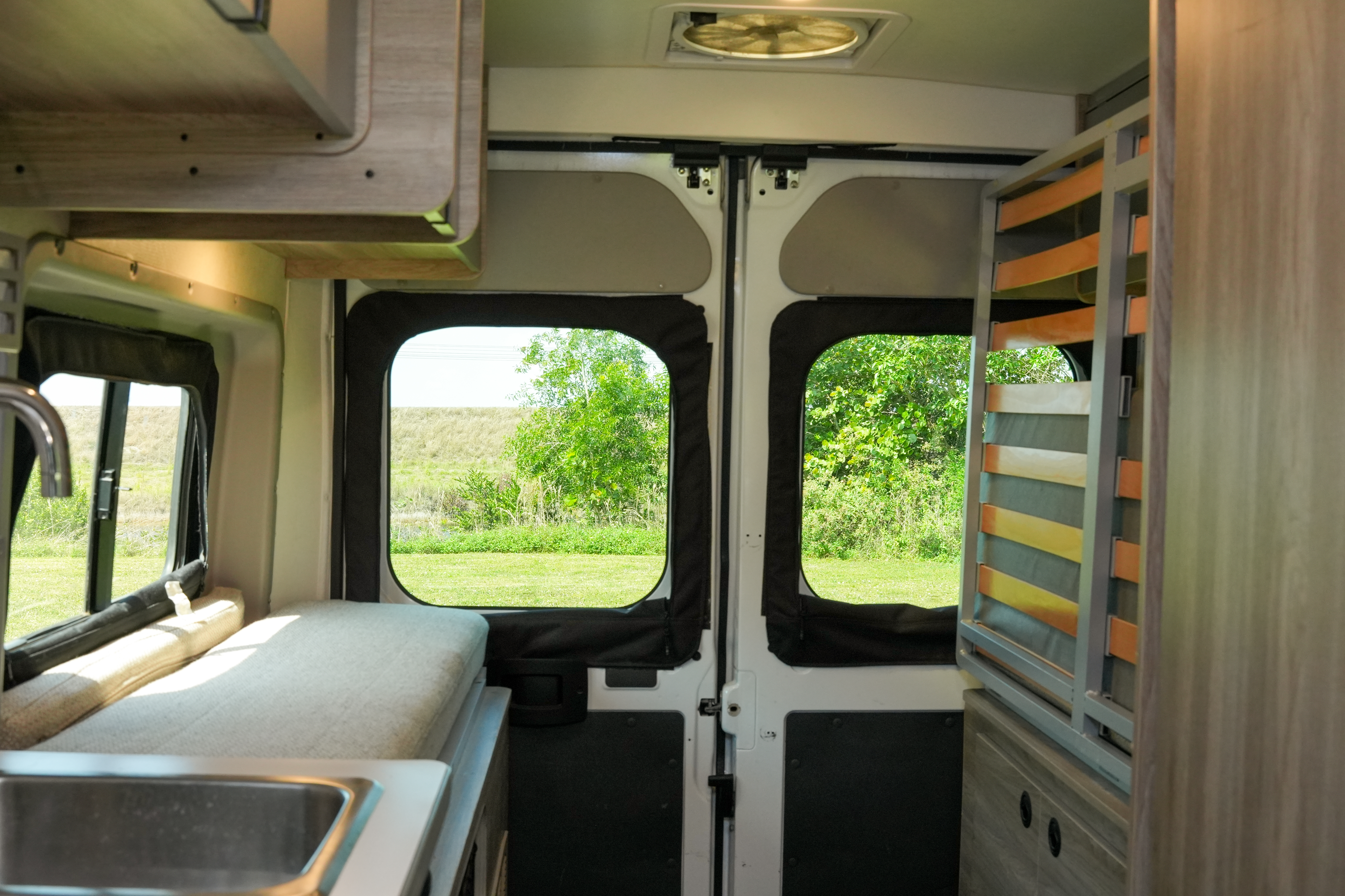 Interior of a campervan with a kitchen sink, foldable bed, and rear doors open to a scenic view.