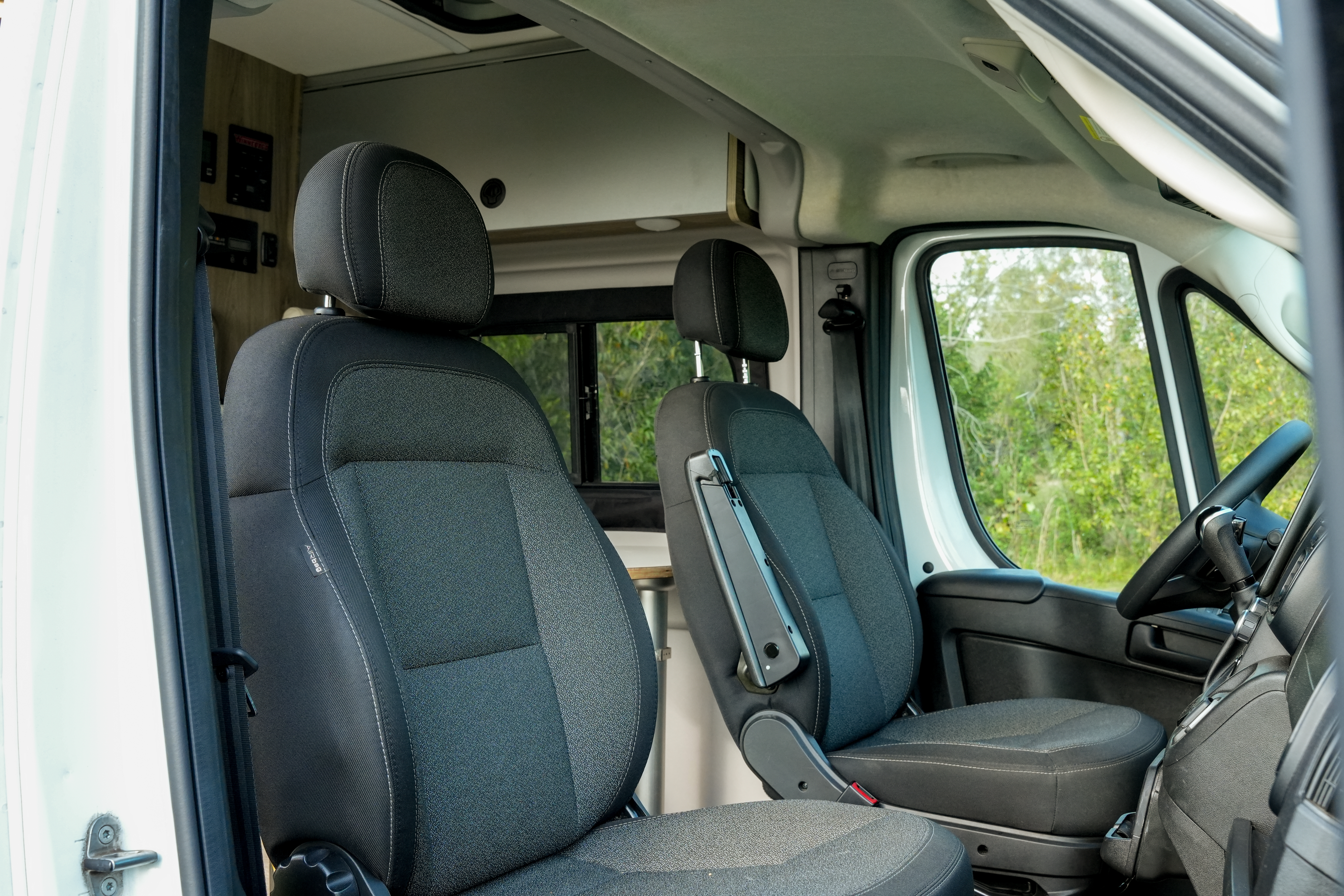 Interior view of a camper van driver and passenger seats with windows and greenery visible outside.