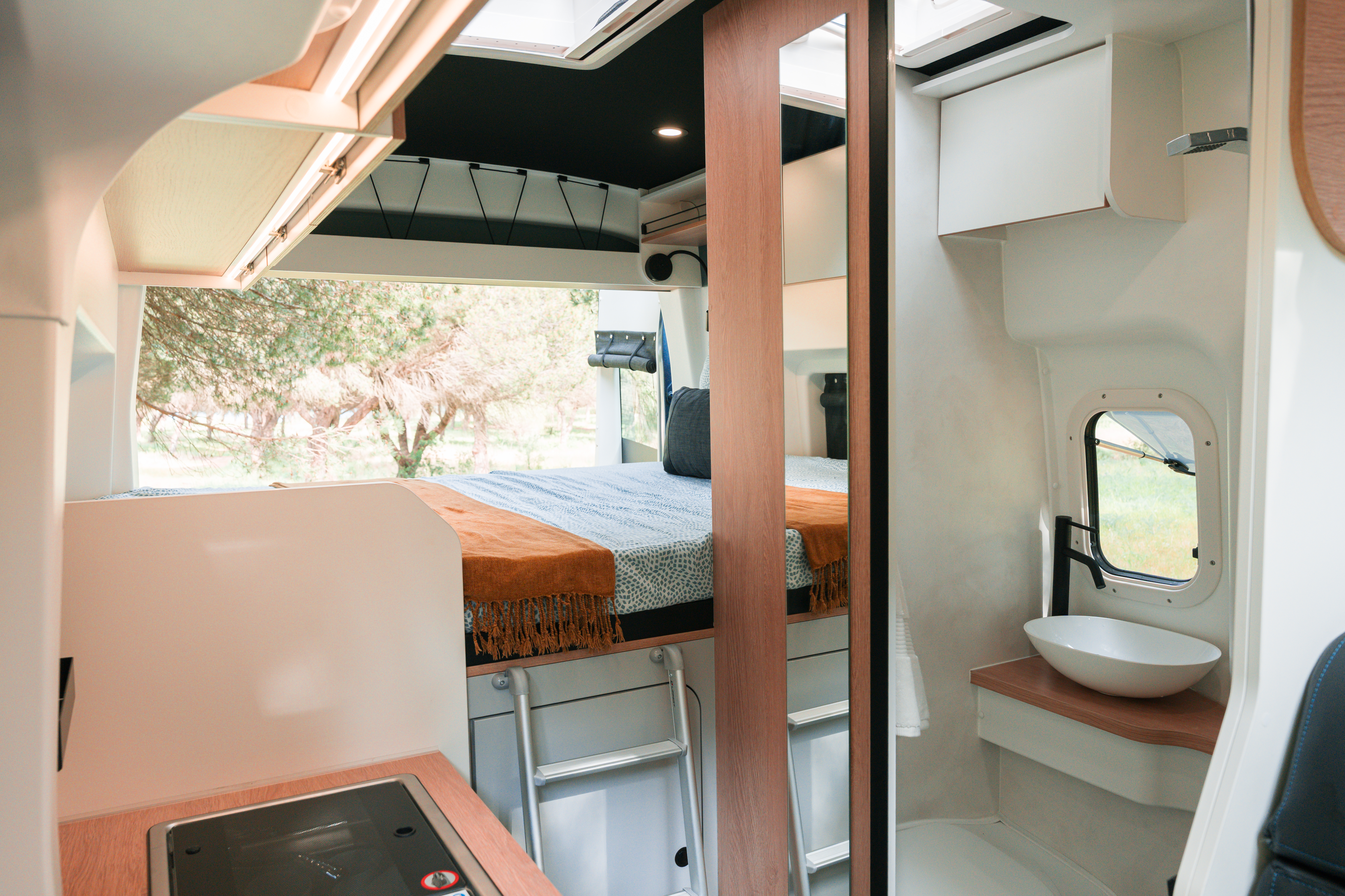 Interno moderno di campervan con letto, cucina compatta, lavandino e finestra, caratterizzato da luce naturale e dettagli in legno.