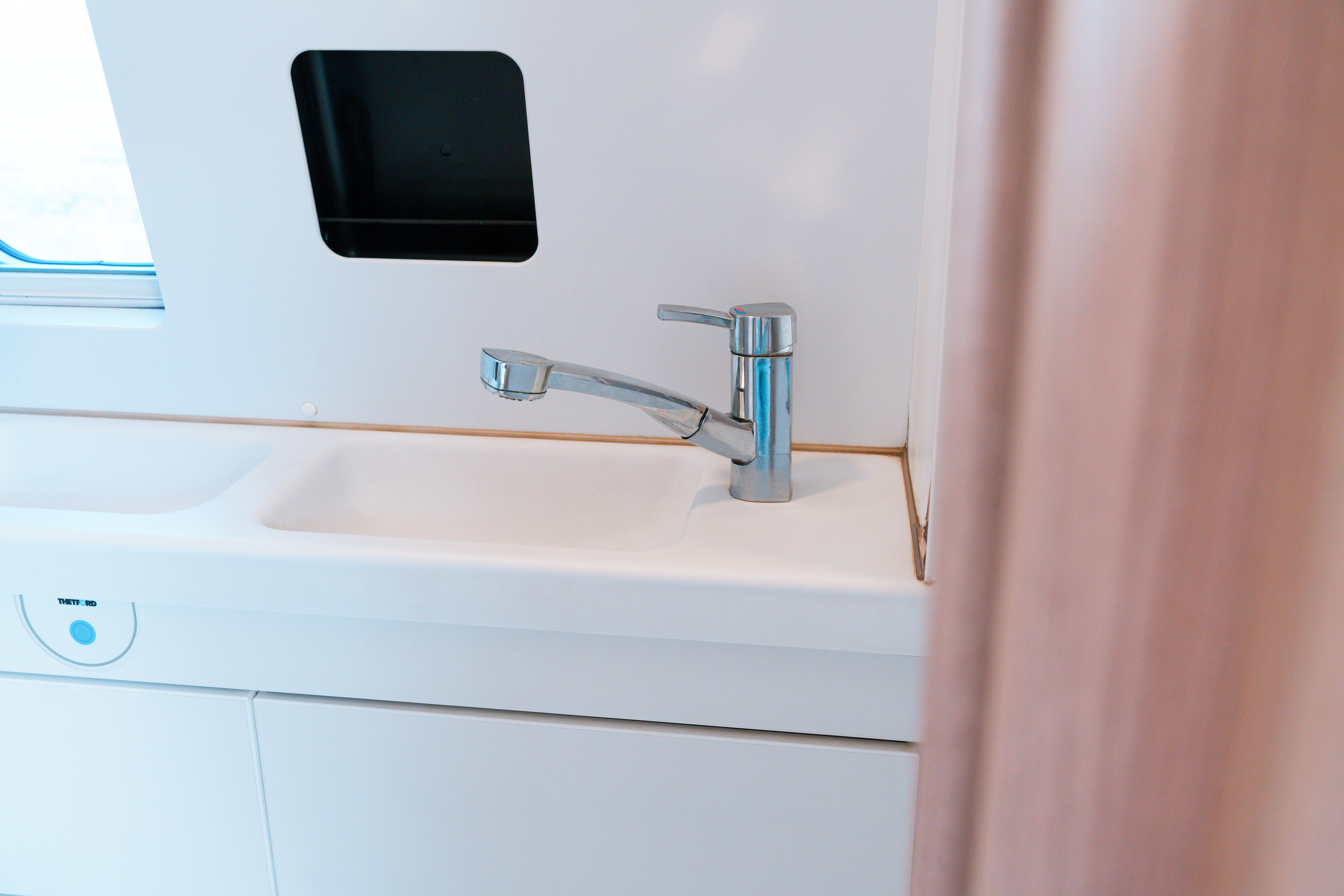 Modern campervan sink with chrome faucet and built-in storage shelf, set against a white countertop and wall.