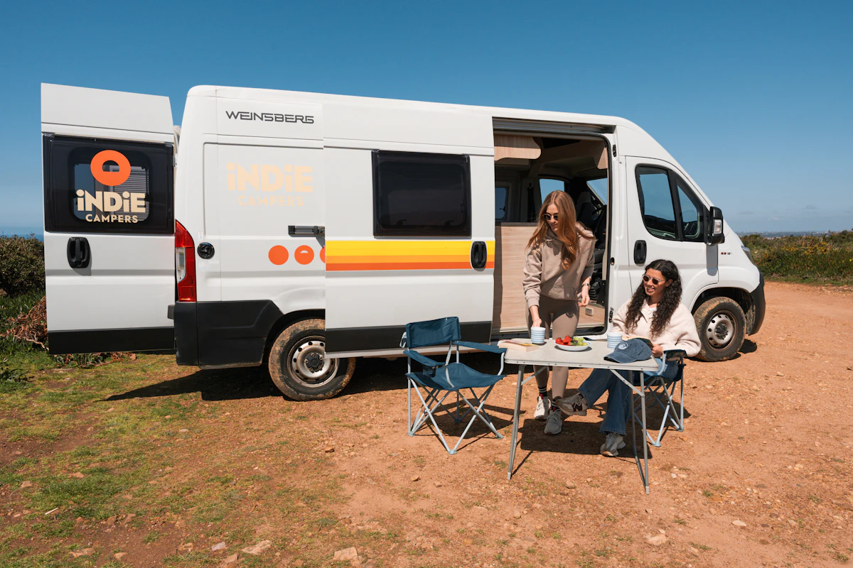 Two women enjoy a meal outside an Indie Campers van parked on a sunny, scenic dirt road.