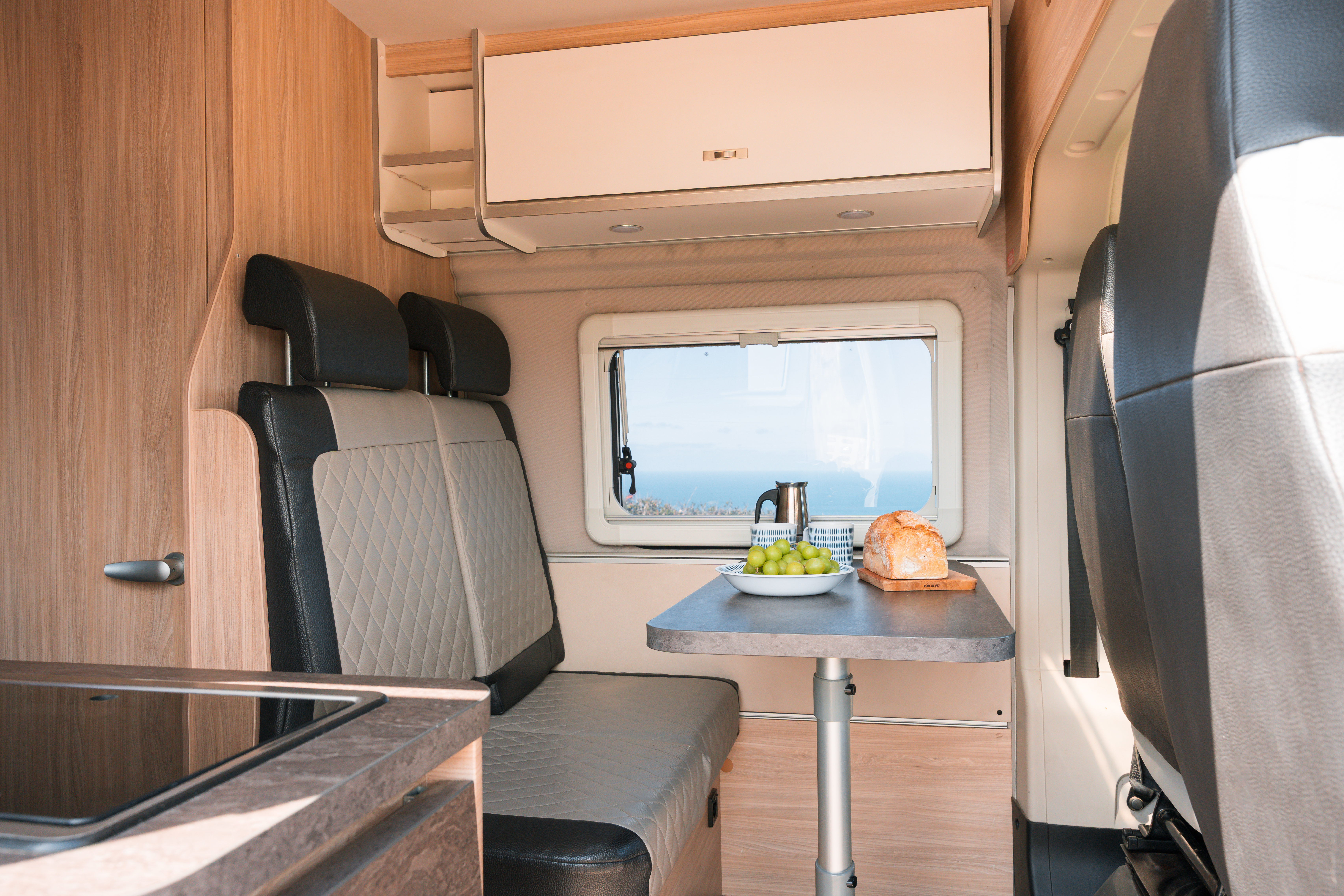 Campervan interior with dining table, bench seats, window view, and food on the table, featuring modern wooden finishes.