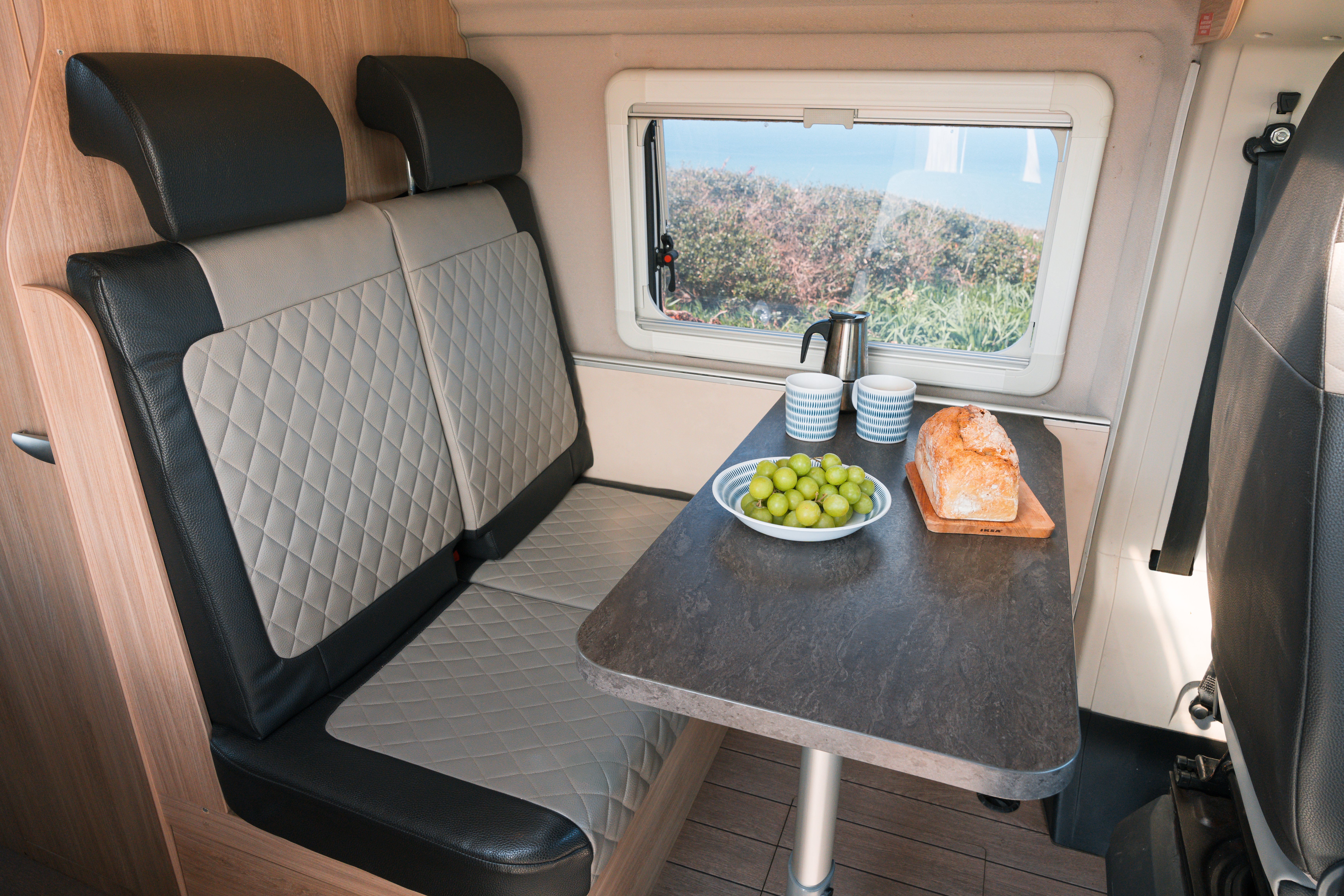 Campervan dining area with cushioned seats, a table set with grapes, bread, cups, and a coffee pot by the window.