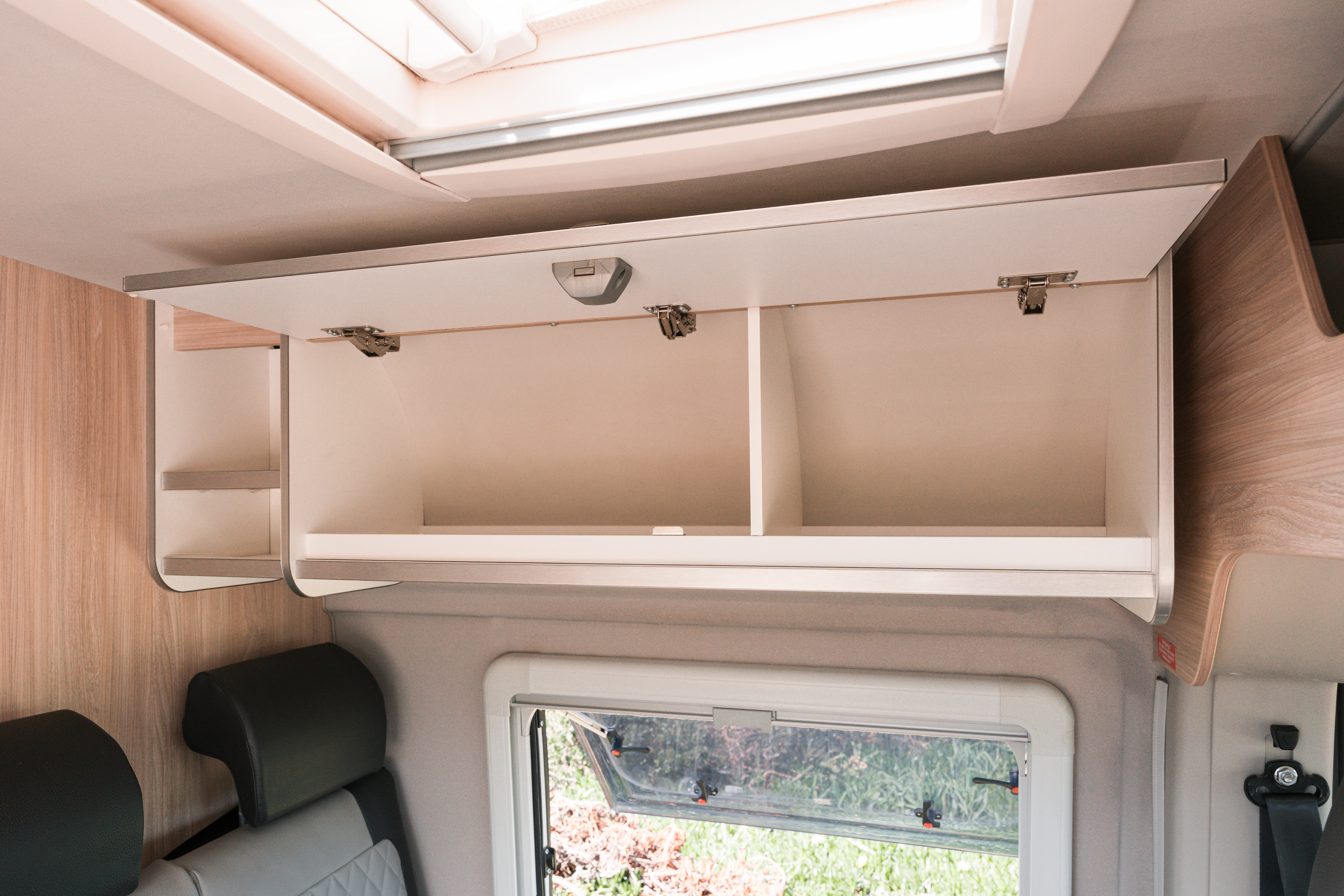 Open overhead storage cabinet with shelves inside a camper van interior, positioned above a window and seating area.
