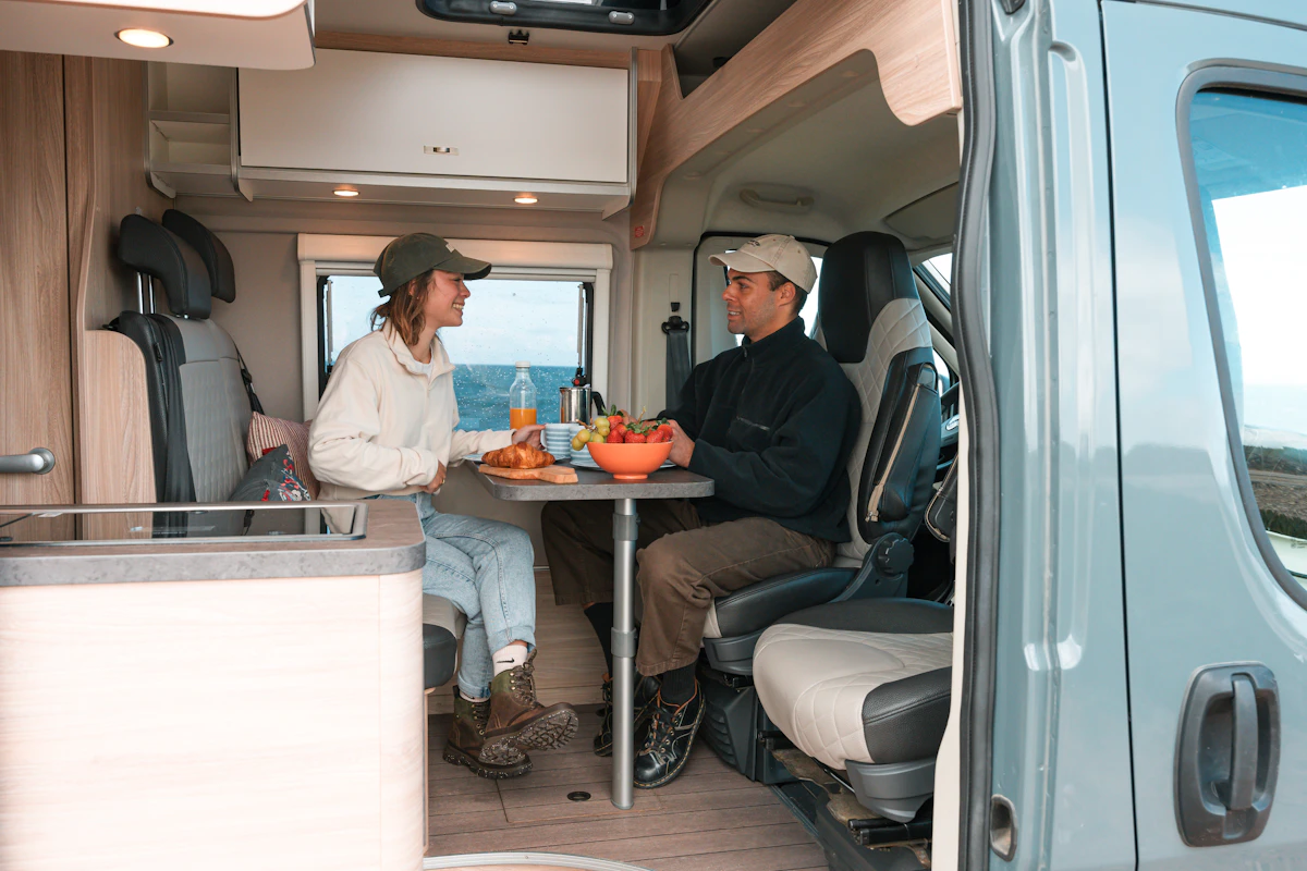 Two people enjoying breakfast at a table inside a modern camper van with outdoor scenery visible through the window.