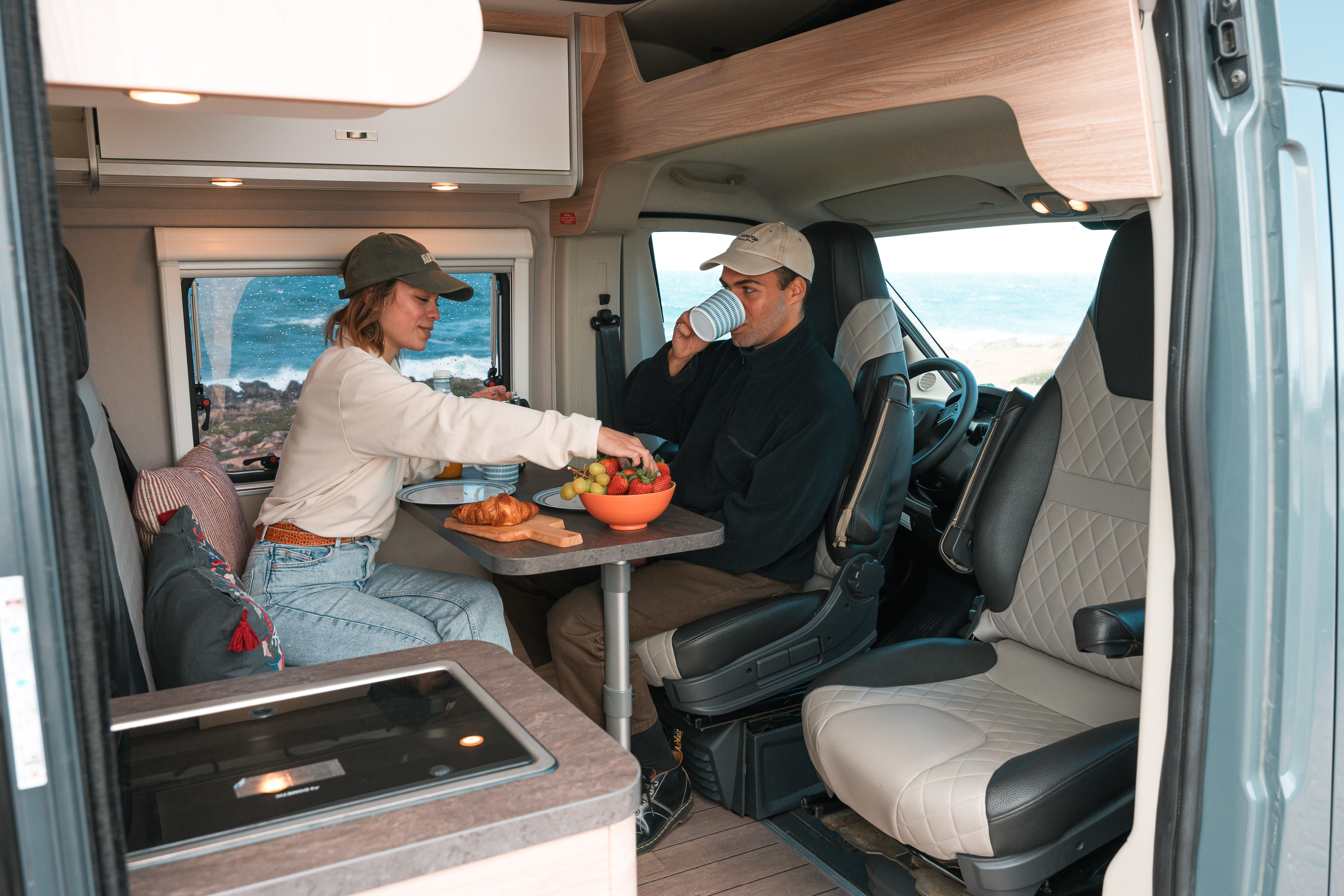 Two people enjoying breakfast inside a modern camper van, with fruit and pastries on the table.