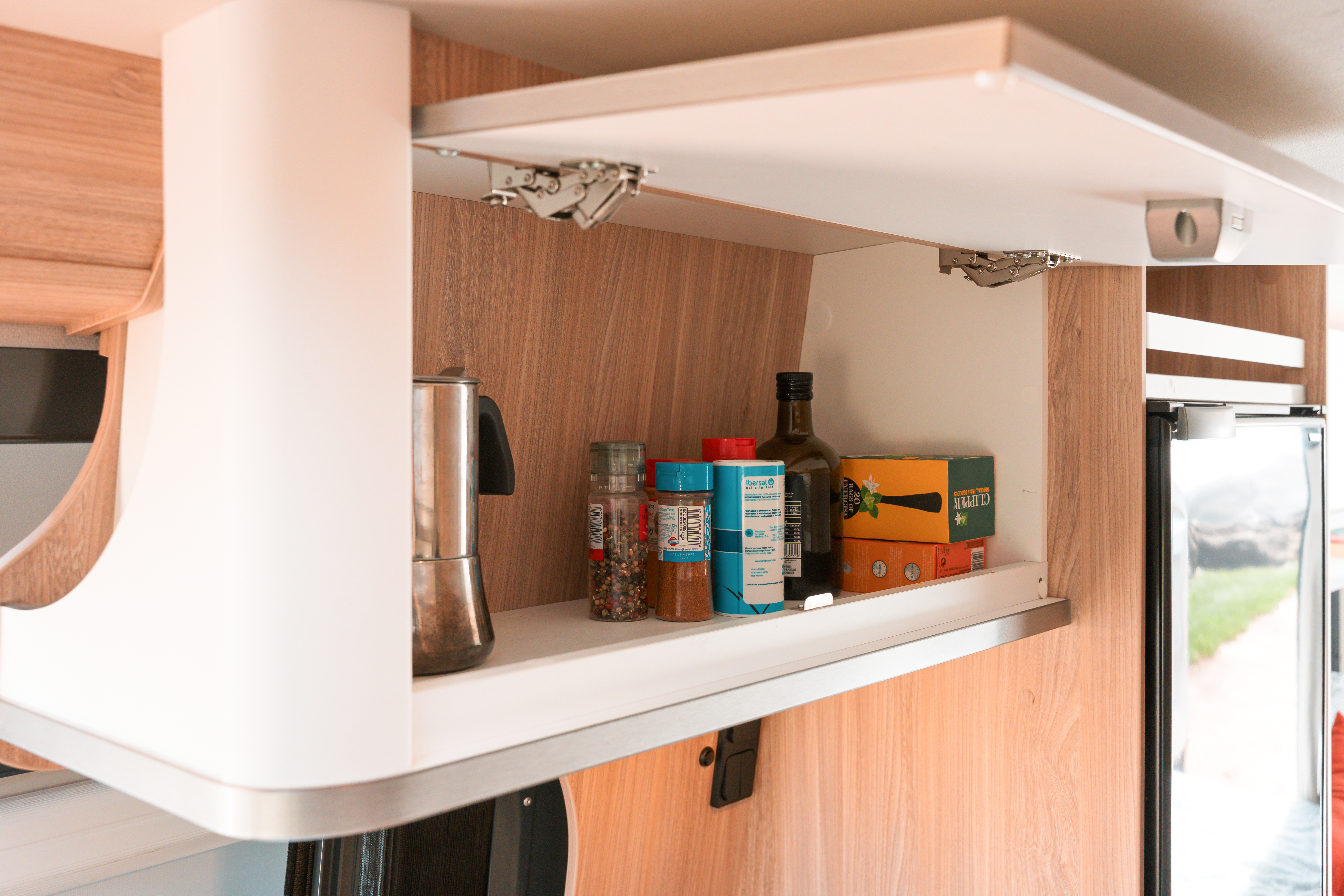 Open campervan kitchen shelf with coffee maker, spice jars, condiments, and food items next to a refrigerator.