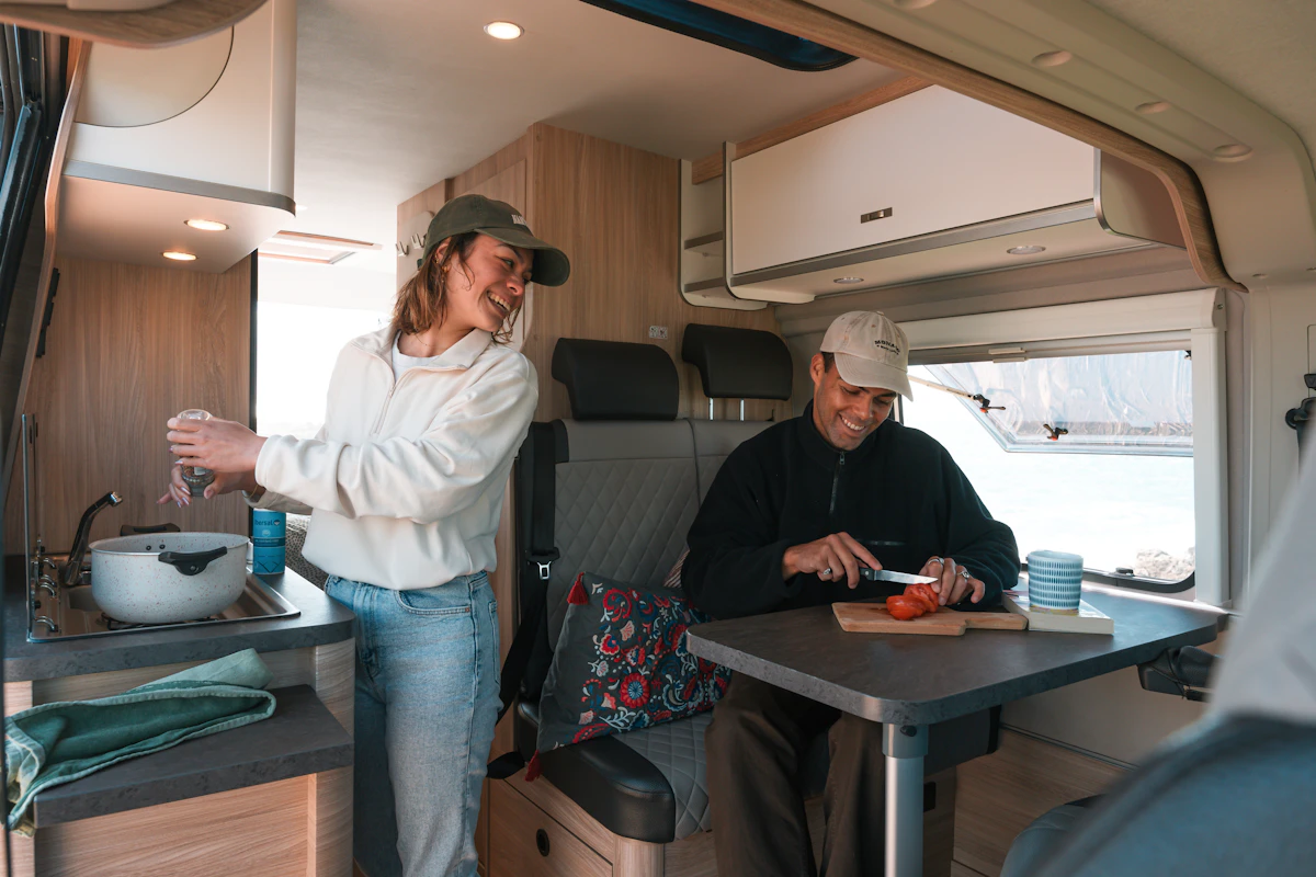 Two people prepare food and smile inside a modern camper van kitchen with a scenic window view.