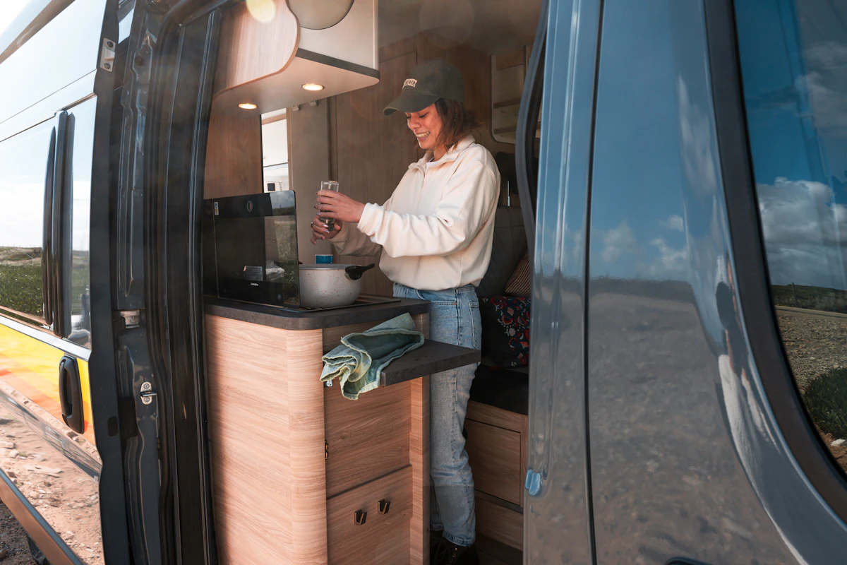 Person preparing a meal inside a camper van with the door open, kitchen area visible, and towels on the counter.