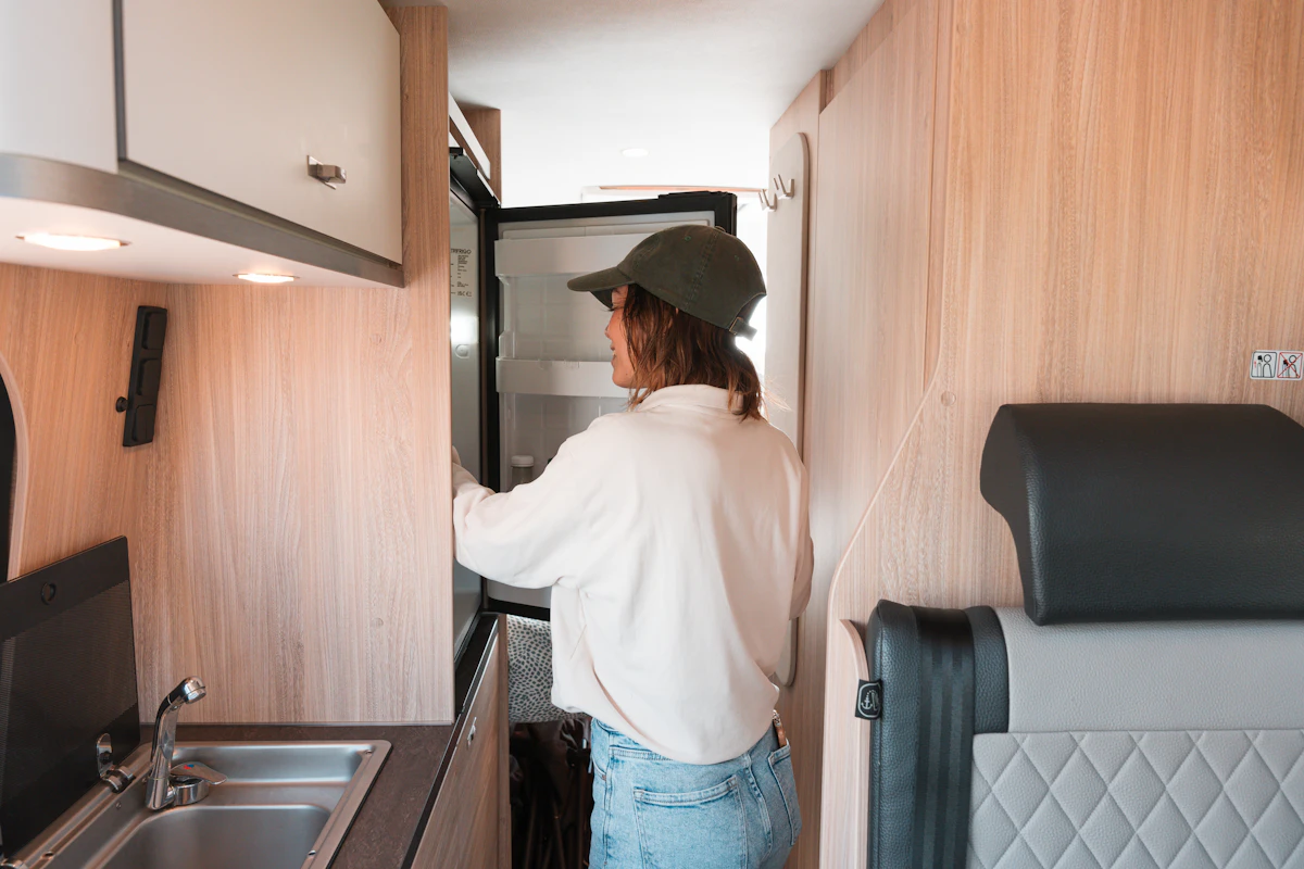 Person standing inside a camper van kitchen, wearing a green cap and opening the refrigerator door.